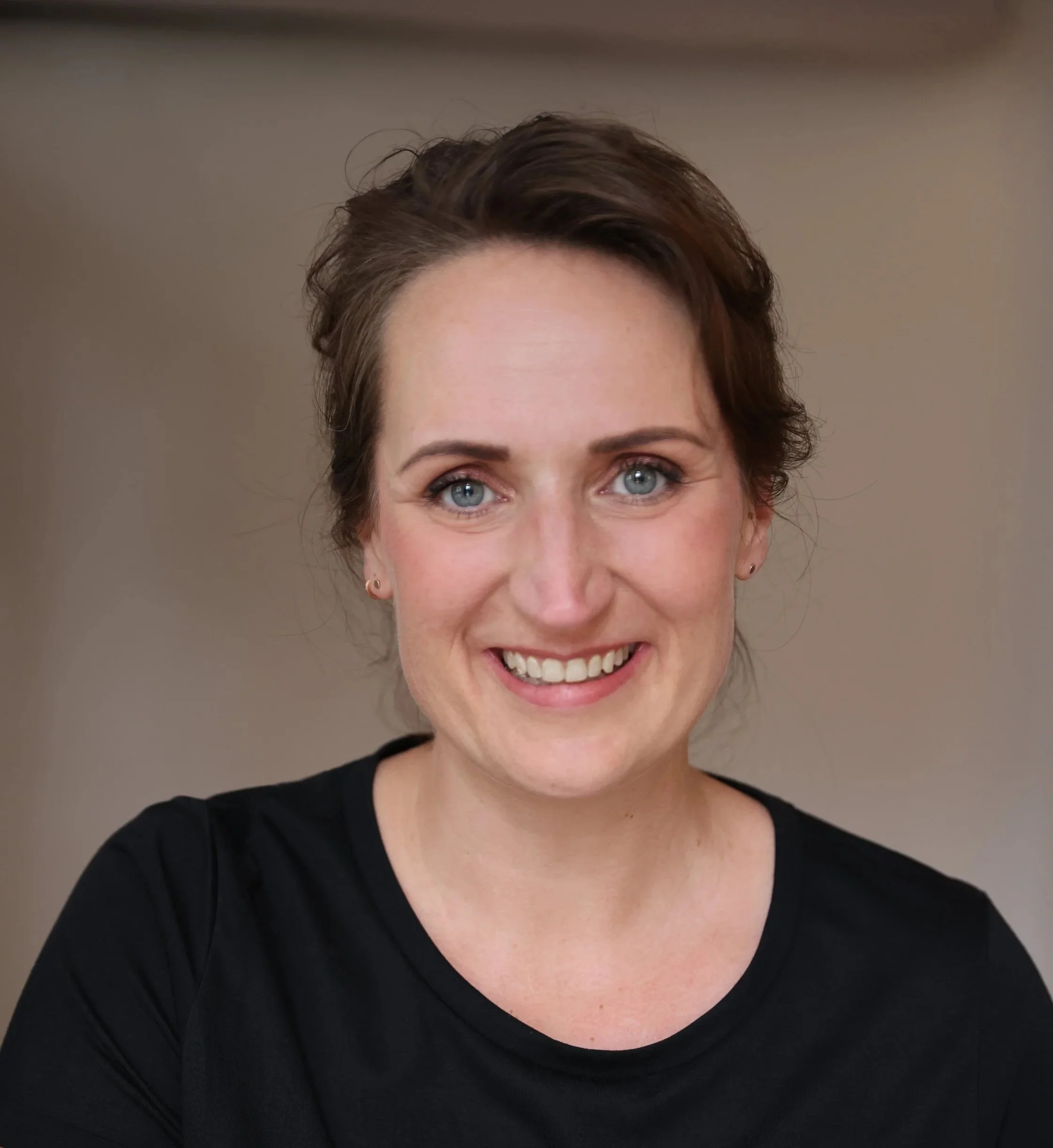 Ein Porträt einer lachenden Frau mit blauen Augen, braunen kurzen Haaren, trägt ein schwarzes Oberteil vor einem neutralen Hintergrund.