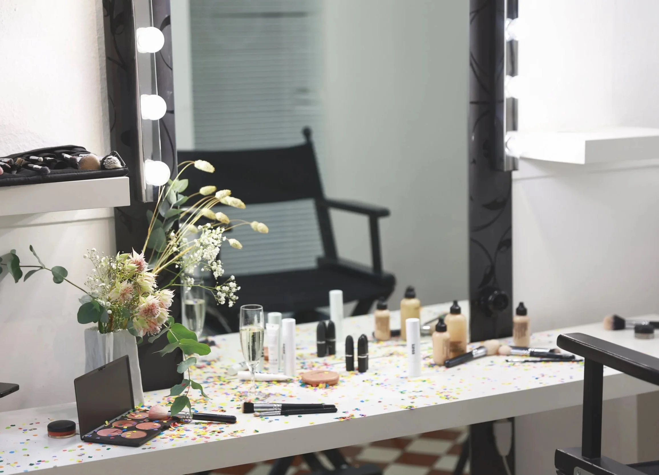 Make-up-Tisch mit verschiedenen Kosmetikprodukten, Make-up-Pinseln, einem Spiegel mit Lichtern, einer Flasche Champagner, einem Blumengesteck und Konfetti auf der Tischoberfläche.