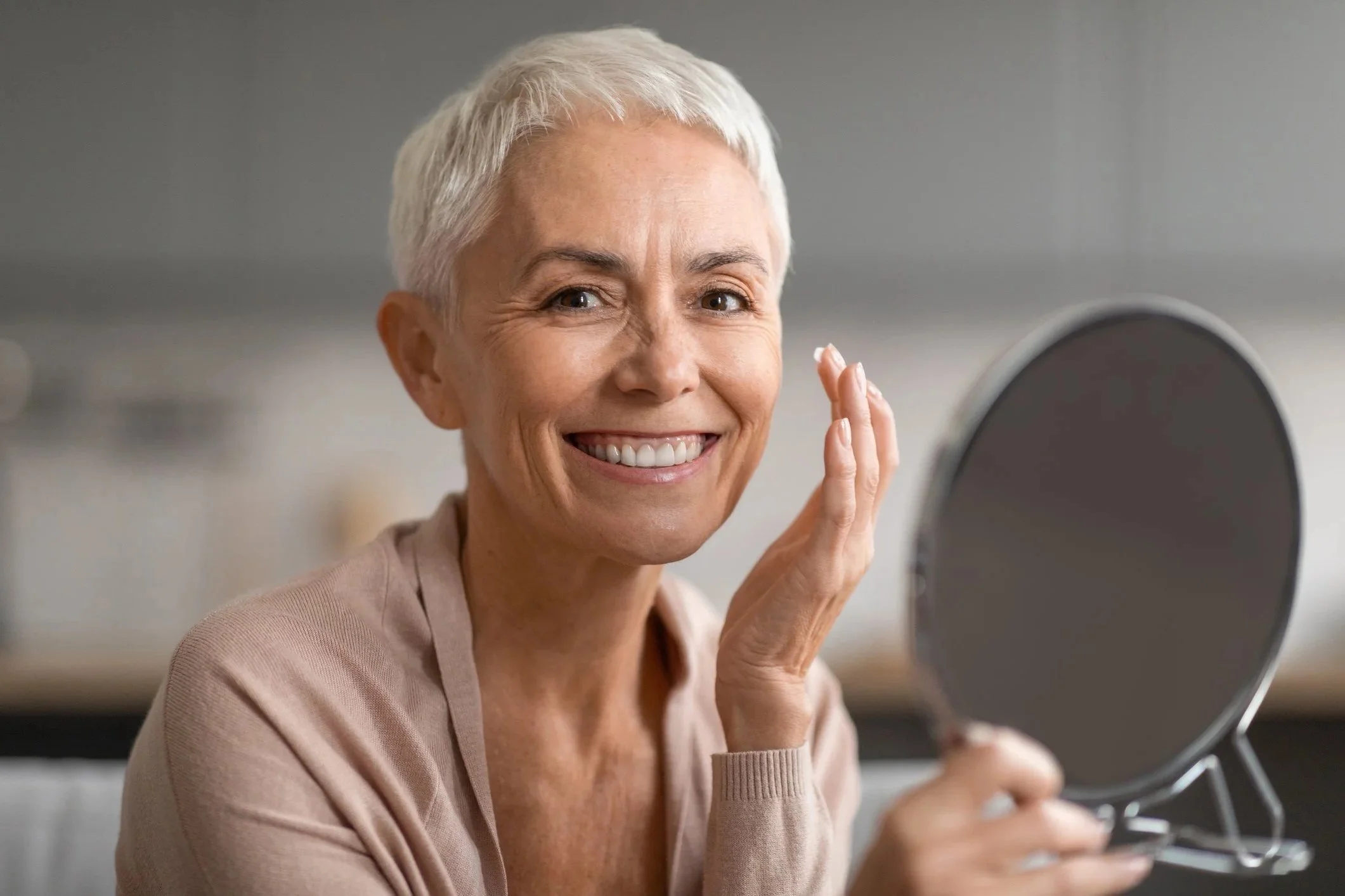 Ältere Frau mit kurz grauem Haar, lächelnd und sich im Spiegel betrachtend.
