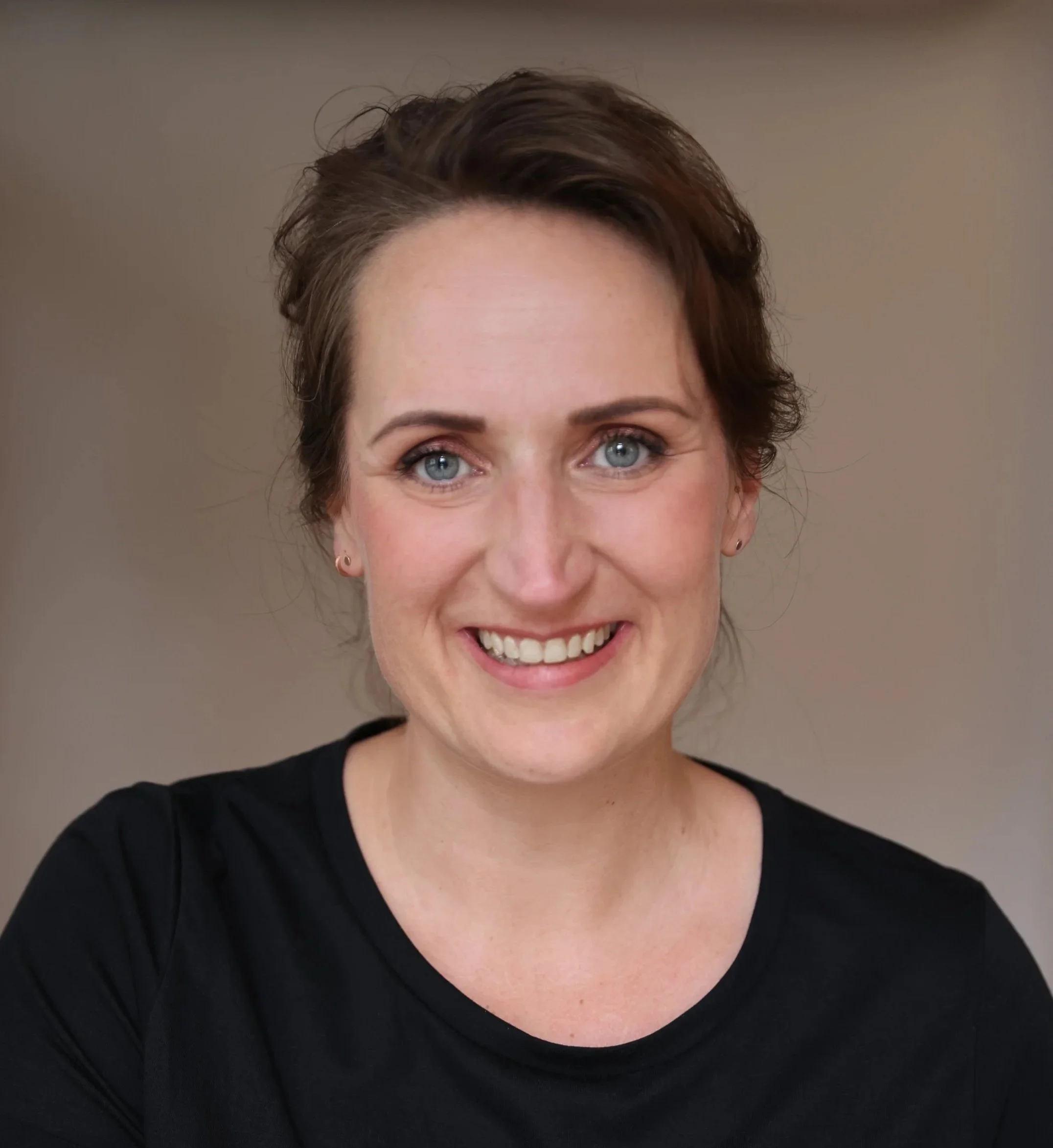 Ein Lächelndes Frau mit blauen Augen, braunen, welligen Haaren in einem schwarzen Oberteil vor einem neutralen Hintergrund.