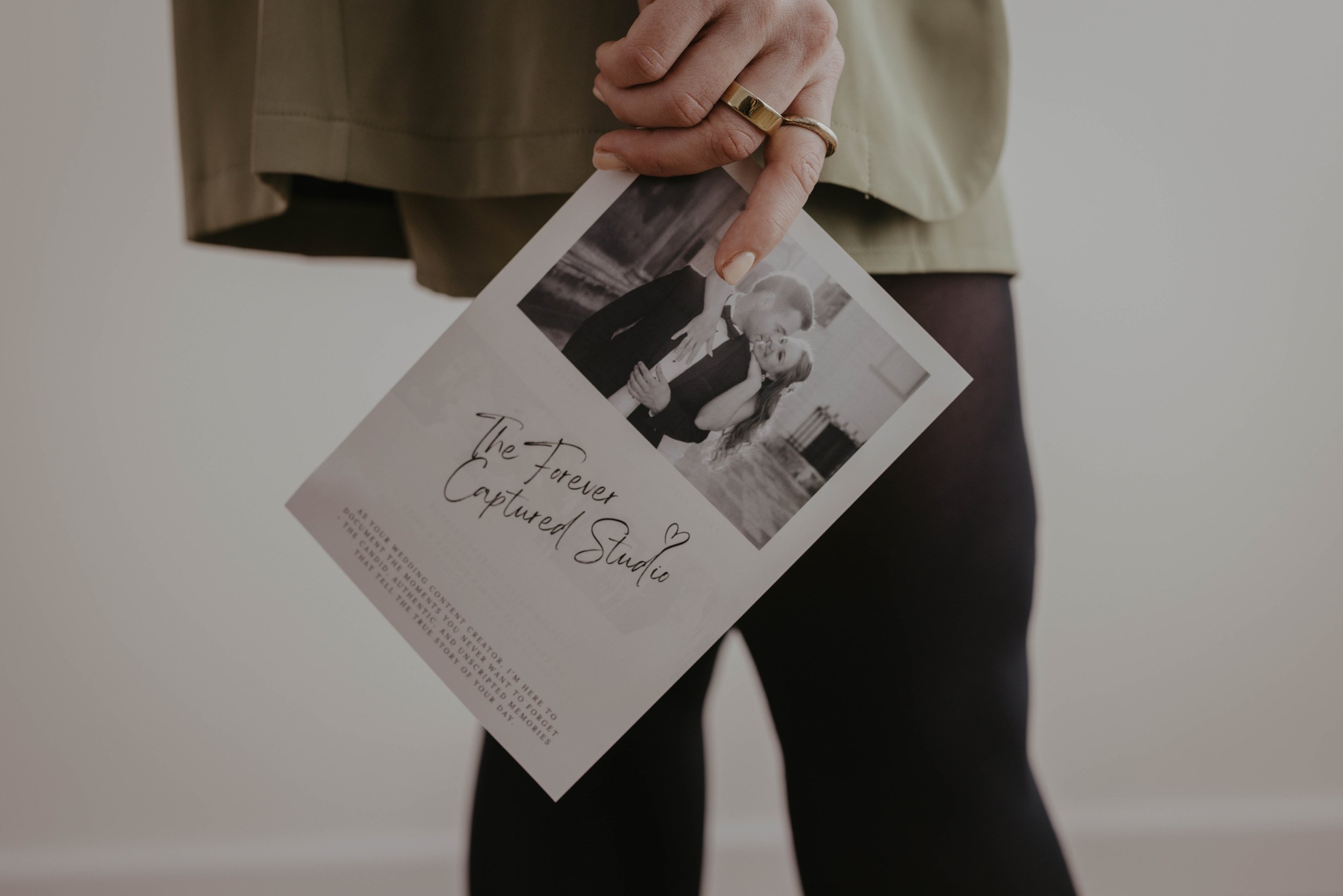 Person holding a printed photograph with a wedding or engagement theme, featuring a couple kissing and the words "The Forever Captured Studio" on the paper.