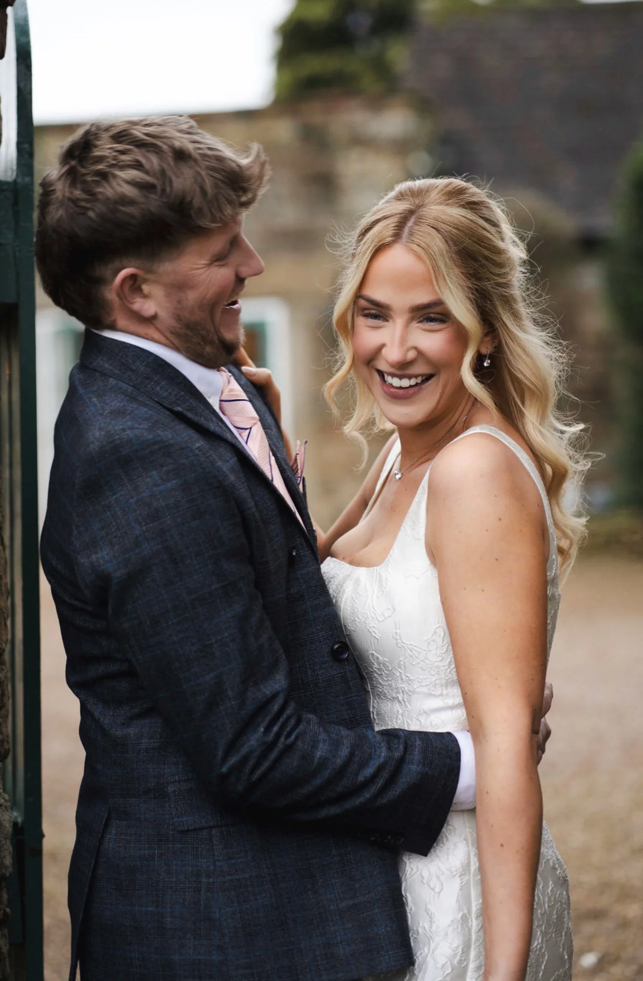 A bride and groom at their wedding reception standing next to a decorated table, with the bride seated on the table and the groom standing beside her, dressed in formal wedding attire, outside at the wedding venue garden