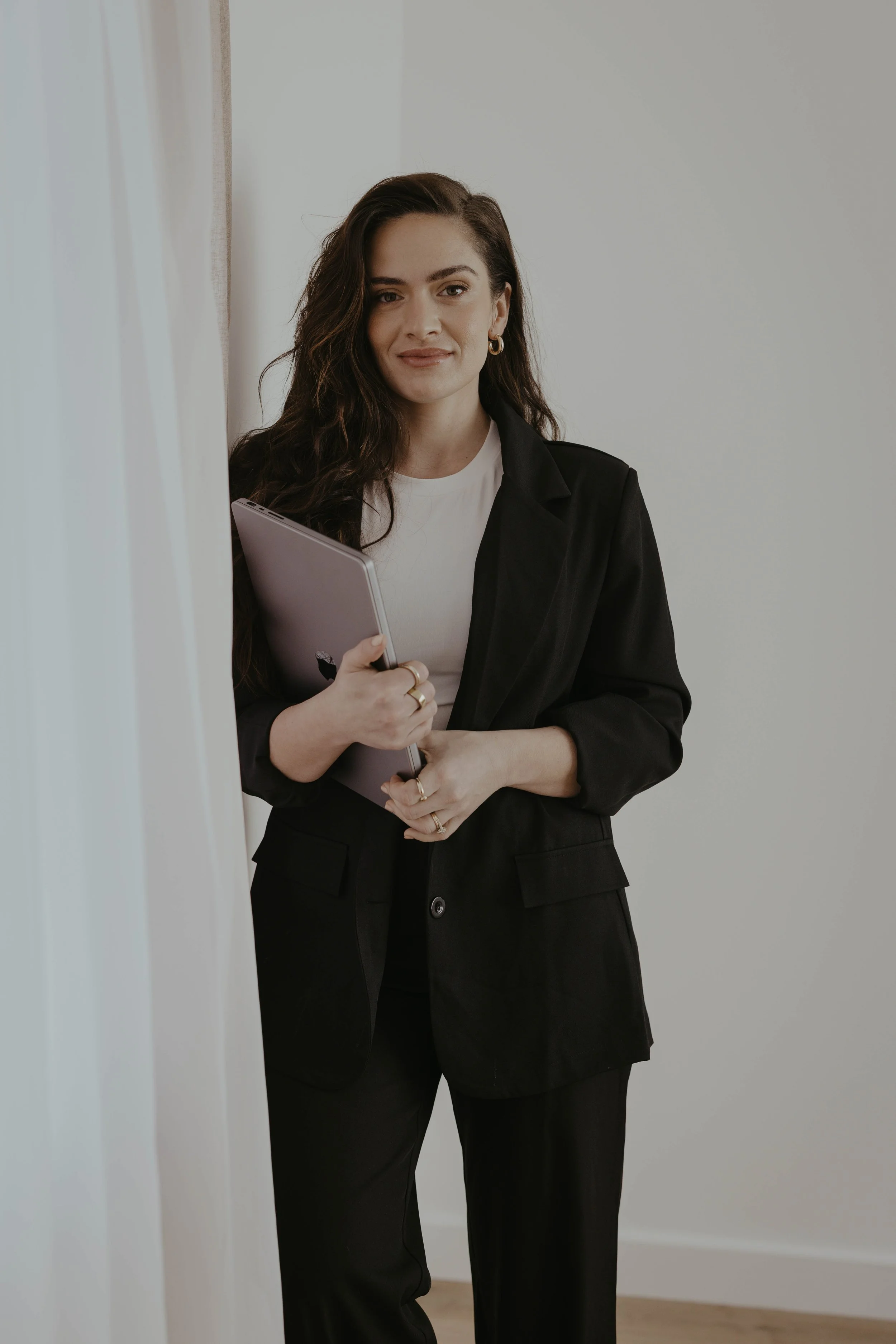 A woman with long dark hair dressed in a black blazer and black pants, holding a closed silver laptop, standing beside a wall with neutral-colored curtains in the background.