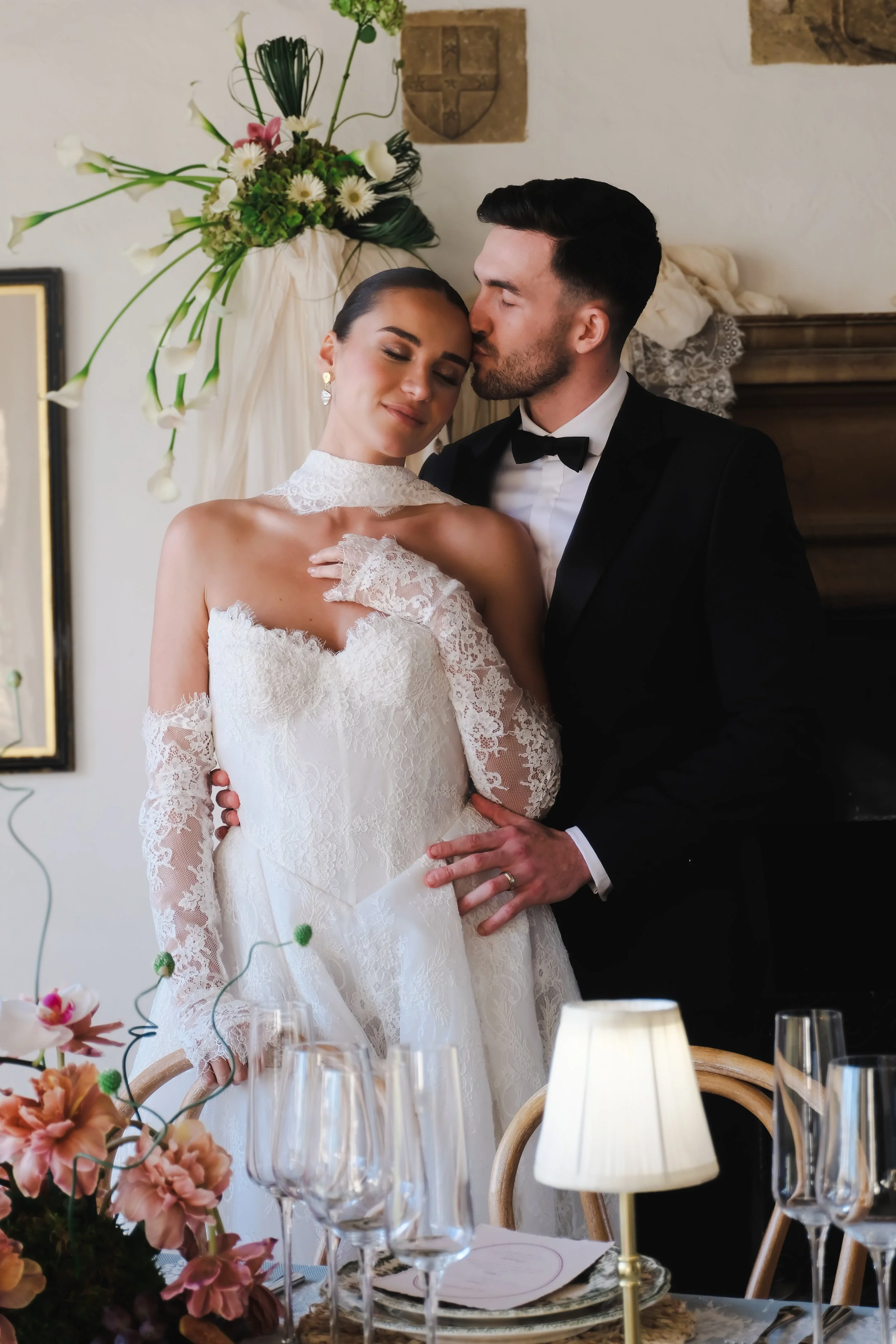 A bride and groom share a tender moment during their wedding reception. The bride wears a lace wedding gown with long lace sleeves and the groom wears a black tuxedo. They are standing close to each other with the groom kissing the bride on her forehead. The setting is decorated with flowers and elegant tableware.