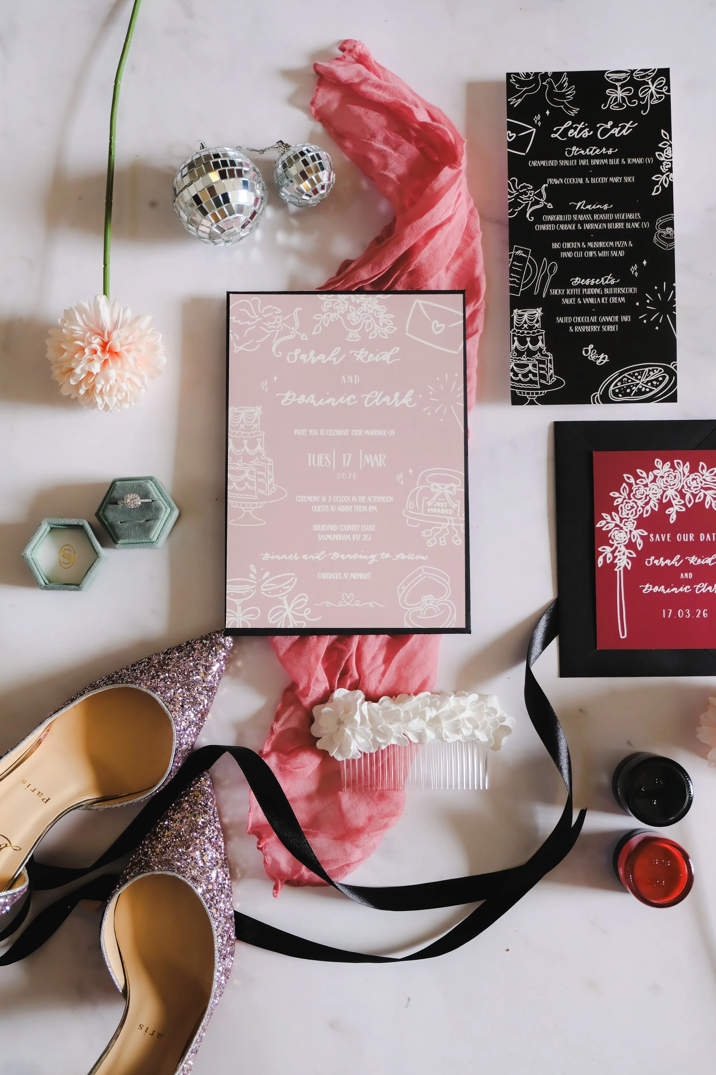 Wedding invitation suite with pink and black backgrounds, pink scarf, pink flower, glittery shoes, glass nail file with white flowers, disco balls, and bottles of nail polish.