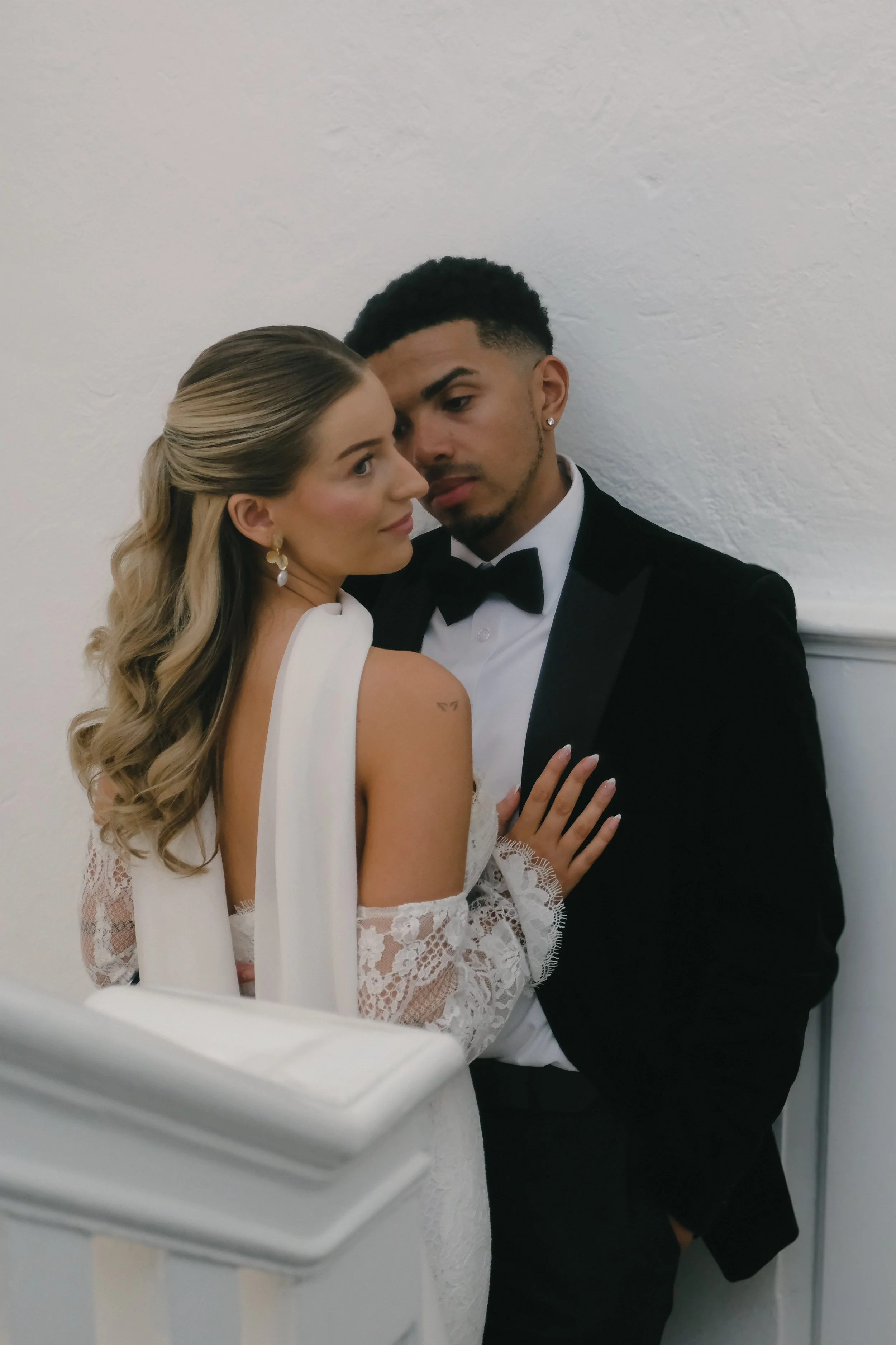 A bride and groom in wedding attire closely embrace against a white wall.