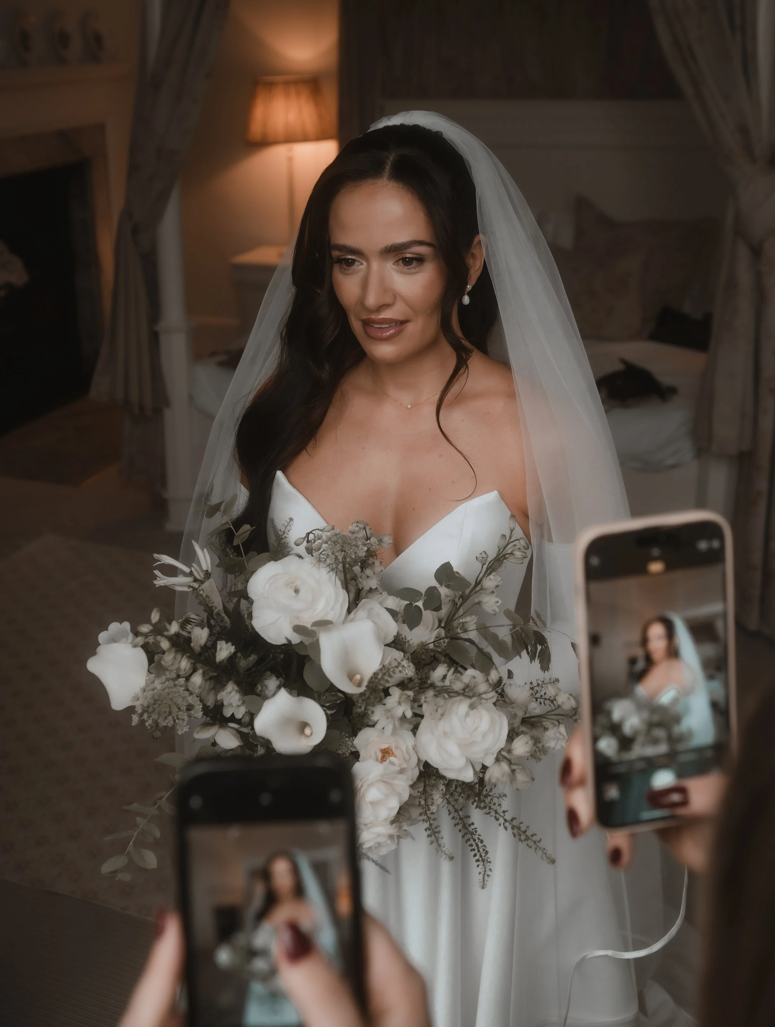 Bride in a wedding dress holding a bouquet of flowers, being photographed by two people with smartphones in a cozy, dimly lit room.