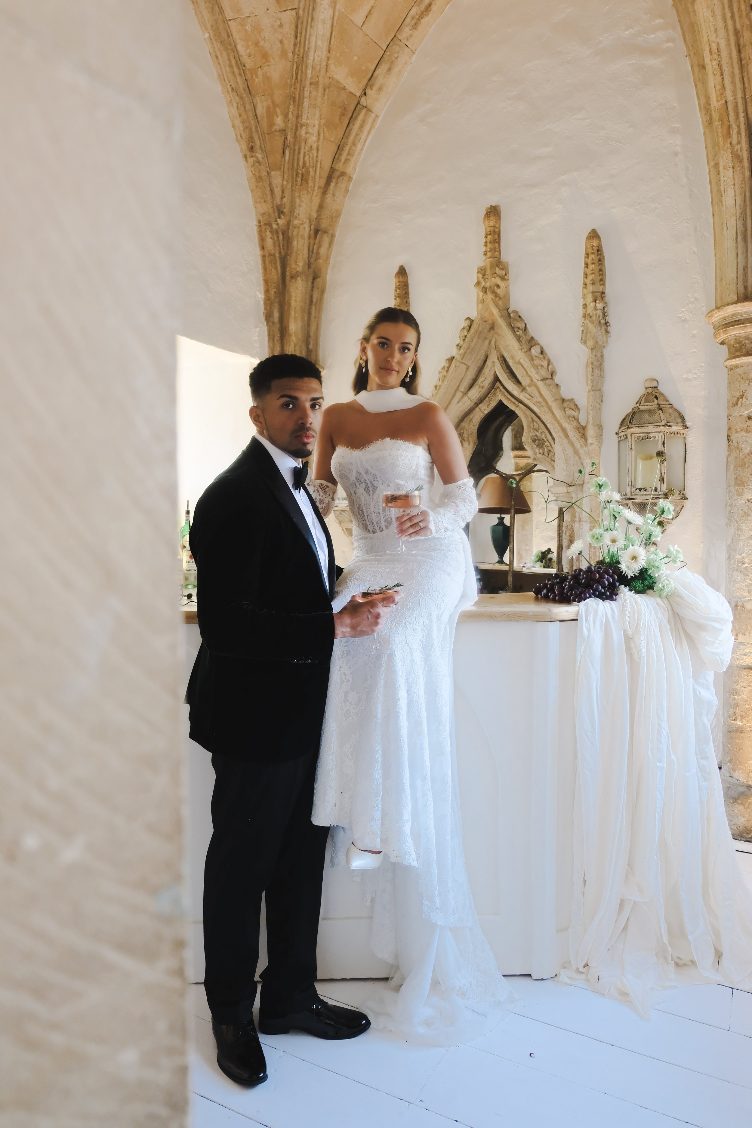 A bride and groom at their wedding reception standing next to a decorated table, with the bride seated on the table and the groom standing beside her, dressed in formal wedding attire, with a backdrop of a stone building interior.