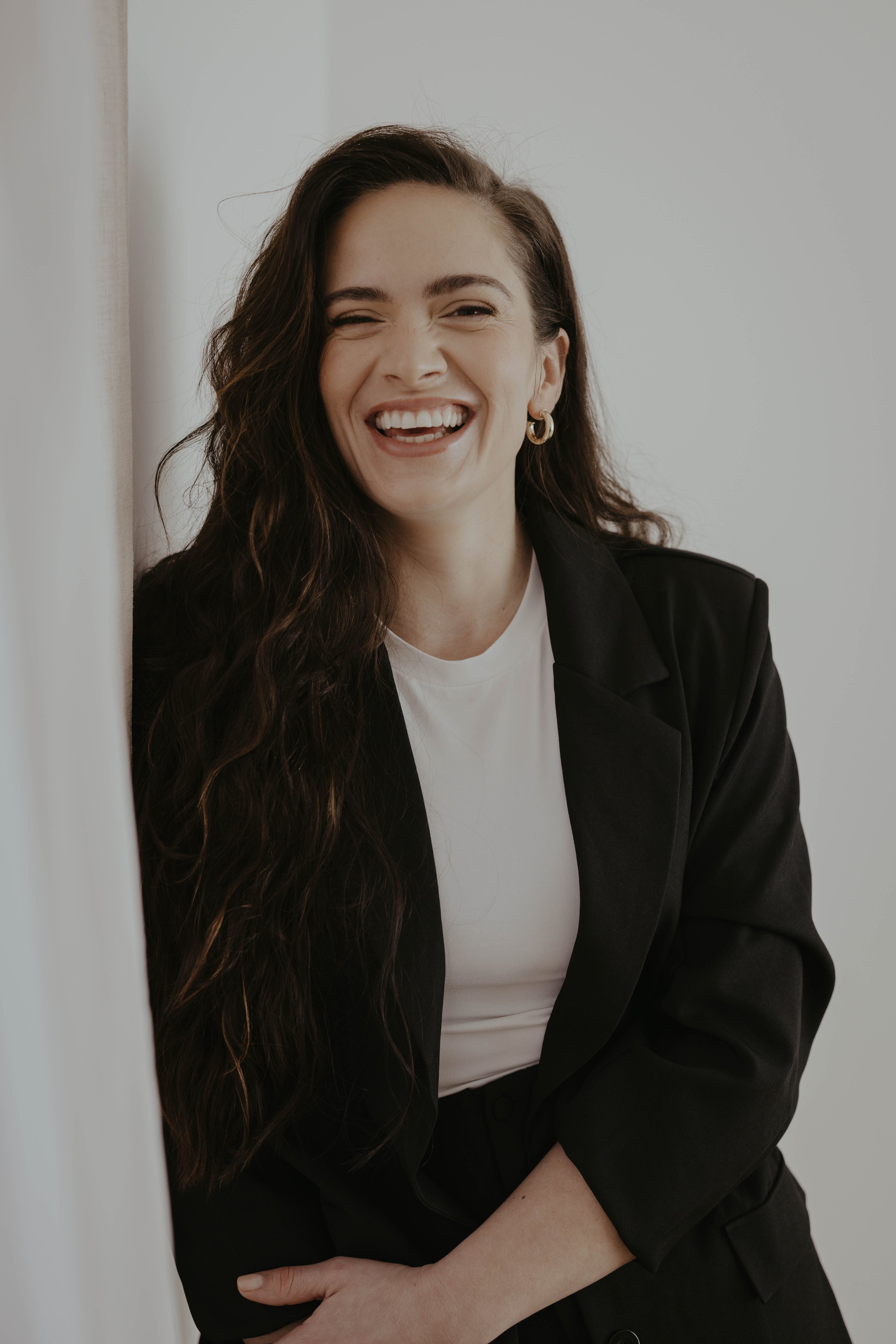 A young woman with long, wavy dark hair wearing a black blazer and white shirt, smiling and laughing while leaning against a wall.