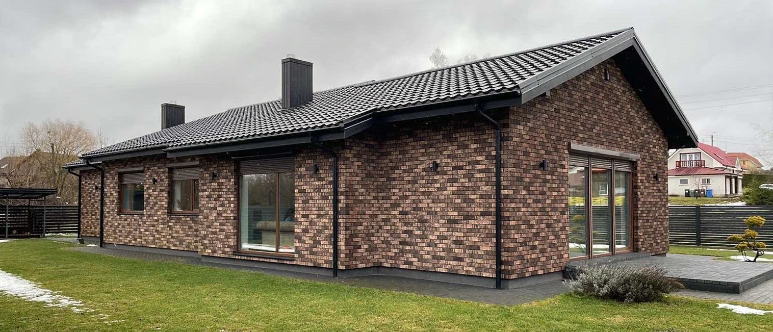 A brick house with a dark gray tiled roof, black trim, and multiple black rain gutters. It has large sliding glass doors and several small windows. The house is surrounded by a well-maintained lawn with some patches of snow and small shrubs, and neig