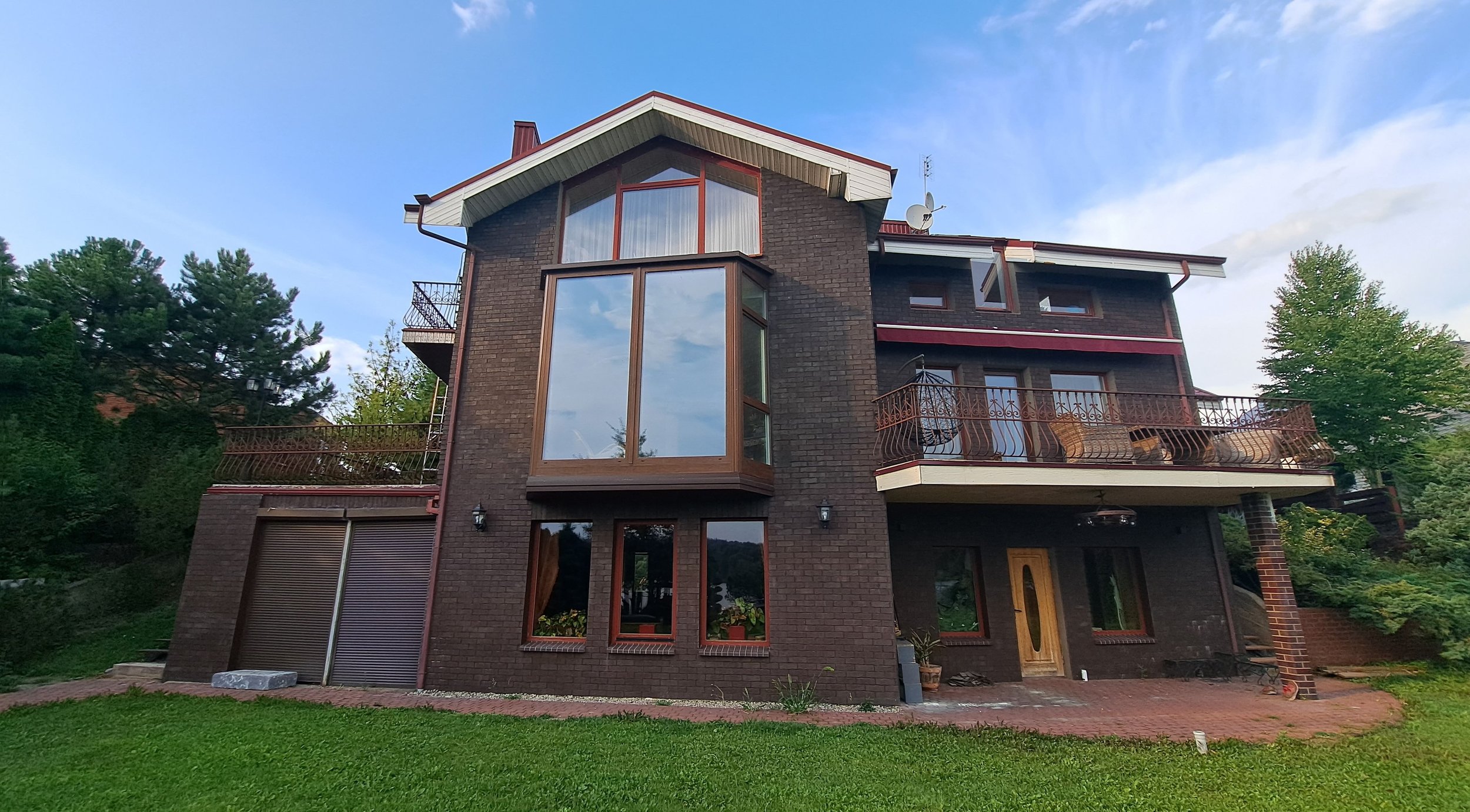 A multi-story brick house with large windows, balconies, and a wooden door, surrounded by green grass and trees under a partly cloudy sky.