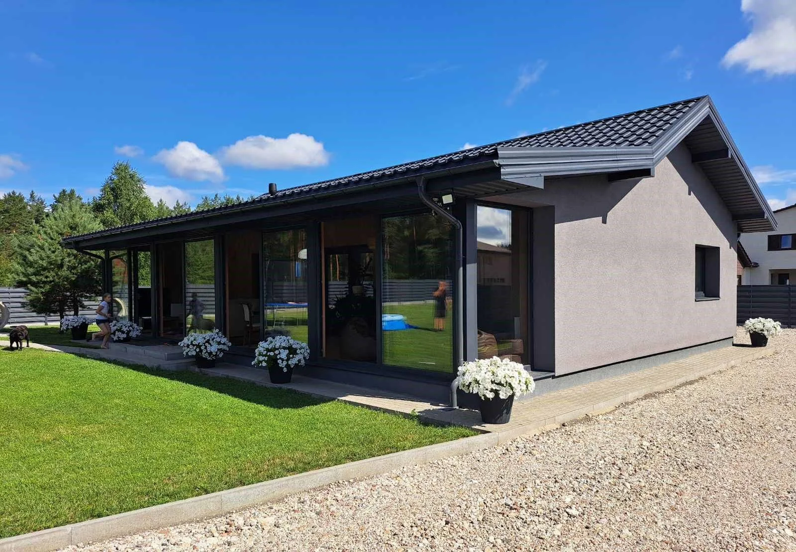 A modern single-story house with a gray roof and beige exterior walls, surrounded by a well-kept lawn and flowerpots with white flowers, under a blue sky with some clouds.