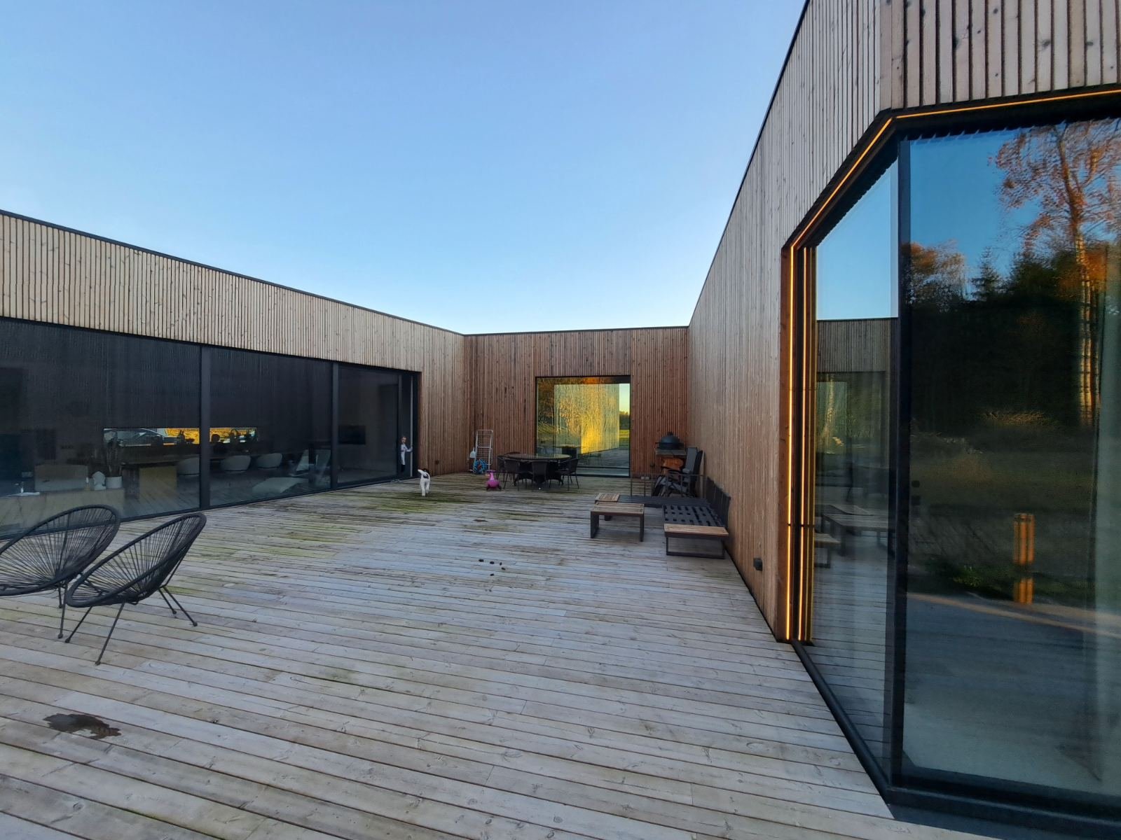 Modern wooden house with a large outdoor deck, black chairs, patio furniture, and glass walls reflecting trees and sky.
