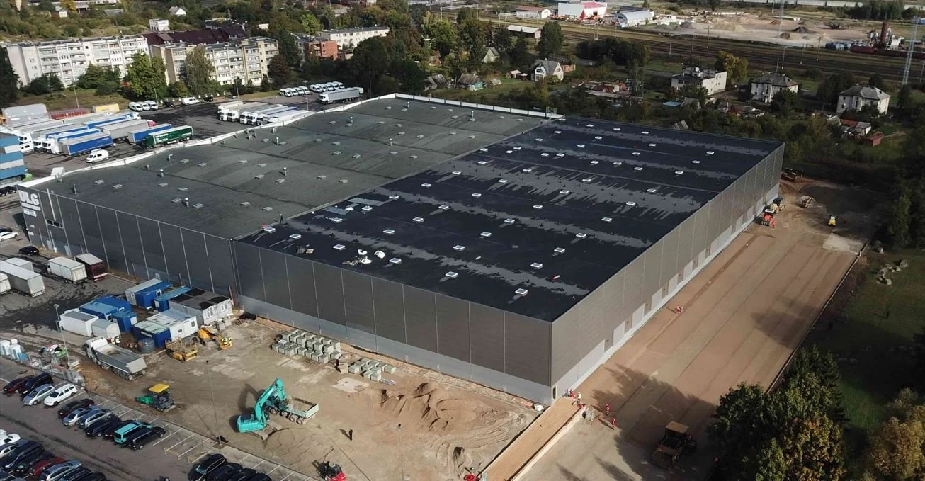 Aerial view of a large industrial warehouse with ongoing construction on one side, surrounded by parking lots and smaller buildings, with residential houses and trees in the background.