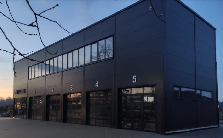A two-story dark gray industrial building with five garage doors numbered 1 through 5. The upper level has a row of windows, and the sky is at dusk.