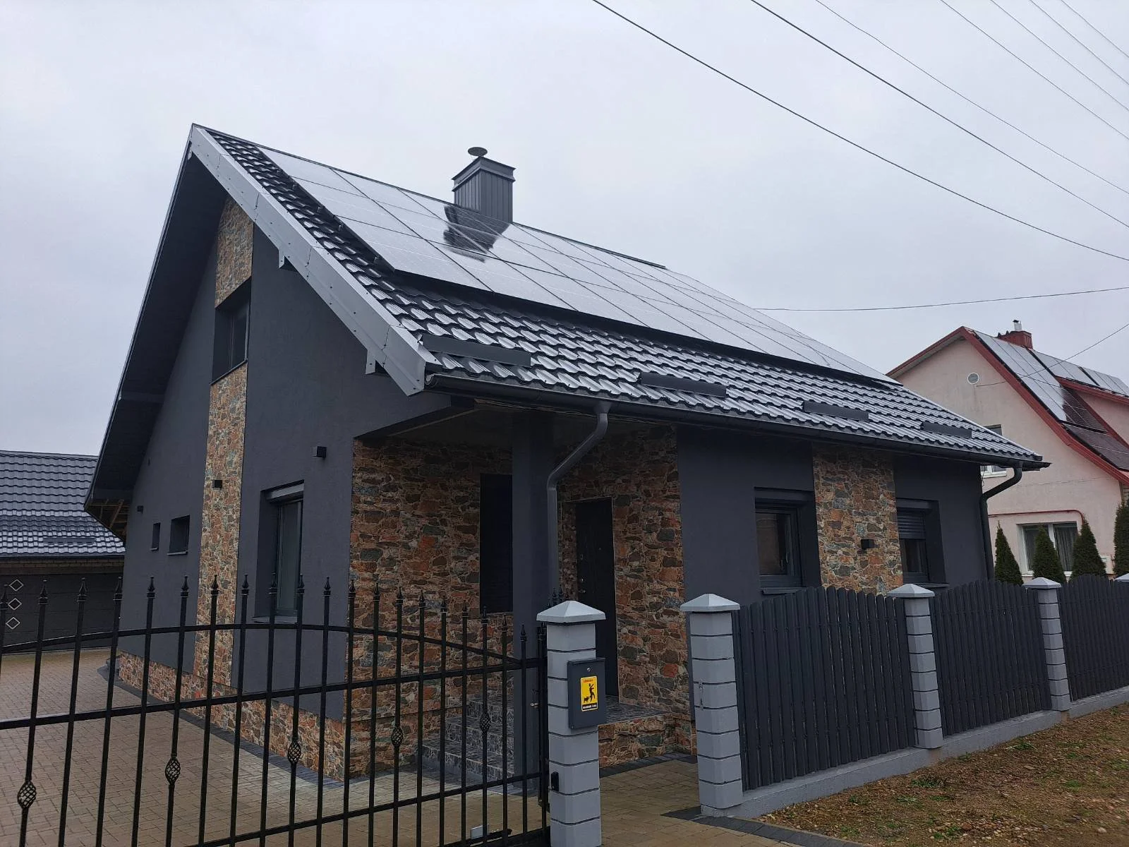 Modern house with dark gray exterior, stone accents, black fence, and solar panels on roof and in background.