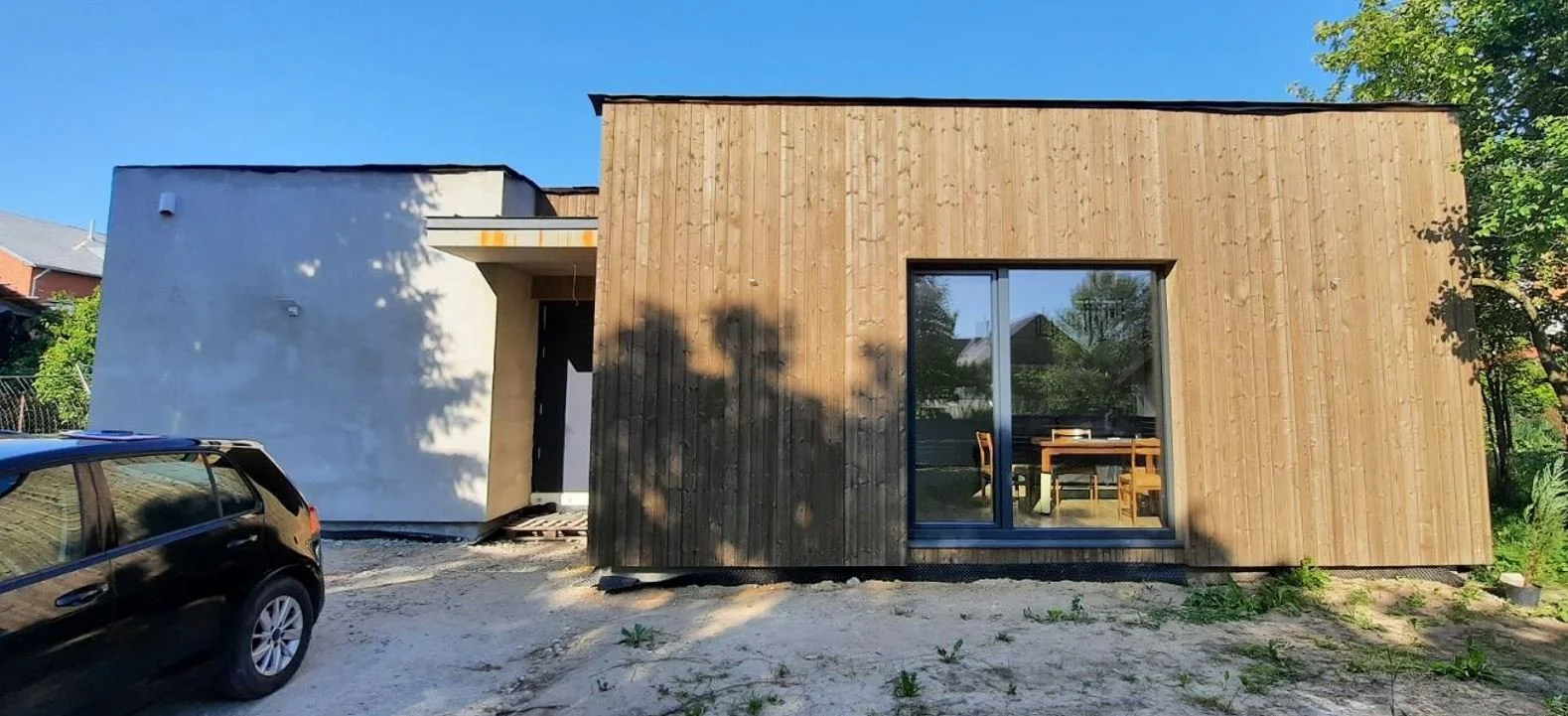 Modern house with a white stucco and wooden exterior, large window, and a car parked in front, under a clear blue sky.