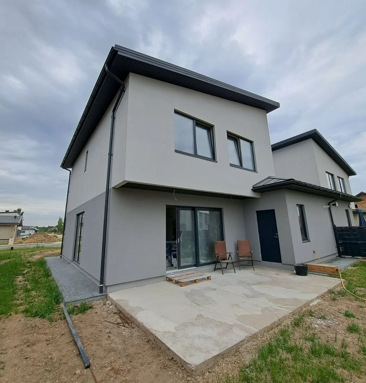 Newly built two-story house with gray exterior walls, black roof, and large windows. Small patio area with outdoor chairs and sliding glass door.