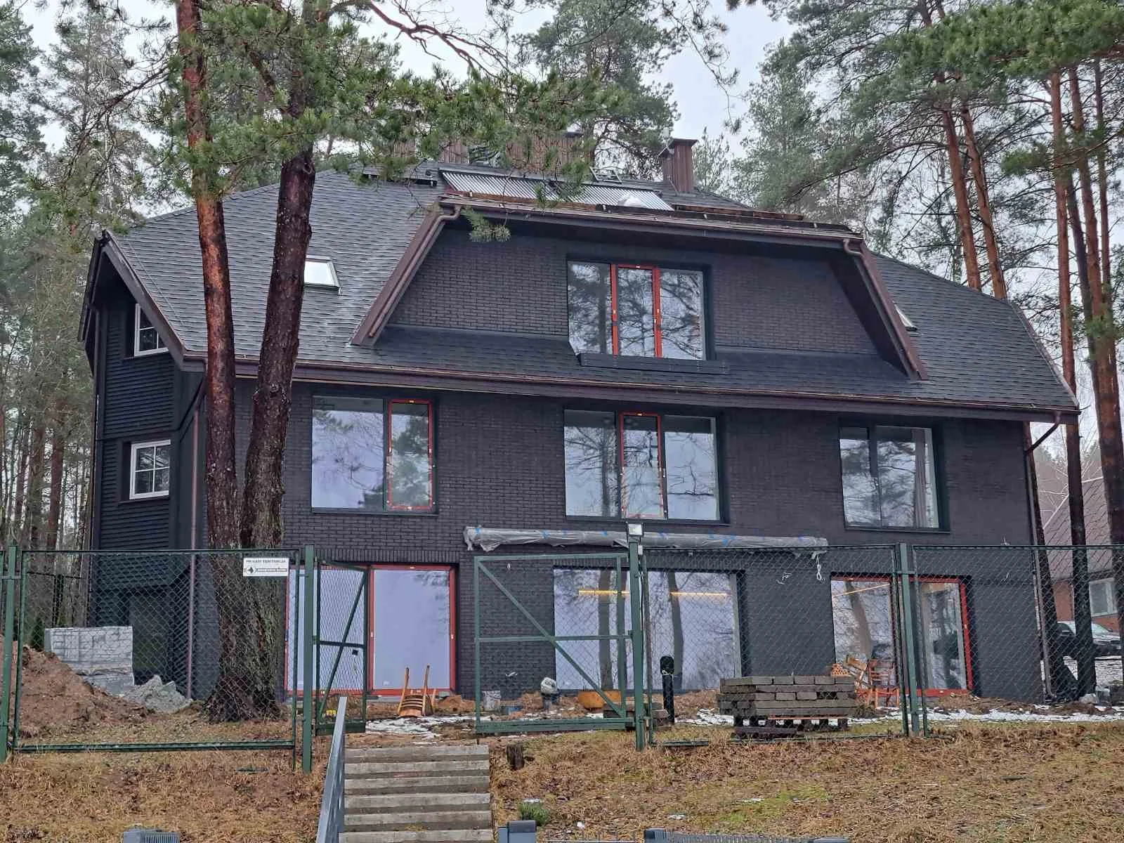A multi-story house under construction, painted black, surrounded by trees, with a fenced yard and construction materials in front.