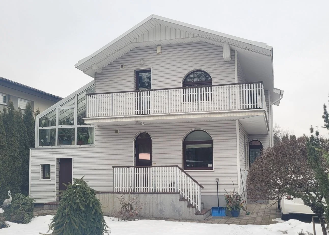A white, three-story house with multiple arched windows, a balcony, and an enclosed sunroom on the left side. The house is surrounded by snow, trees, and shrubs.