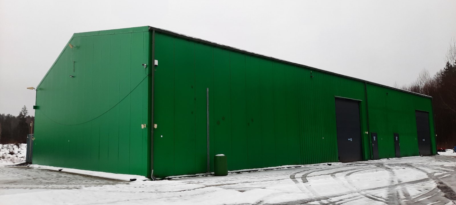 A large green industrial or storage building with black doors, set in a snowy landscape.