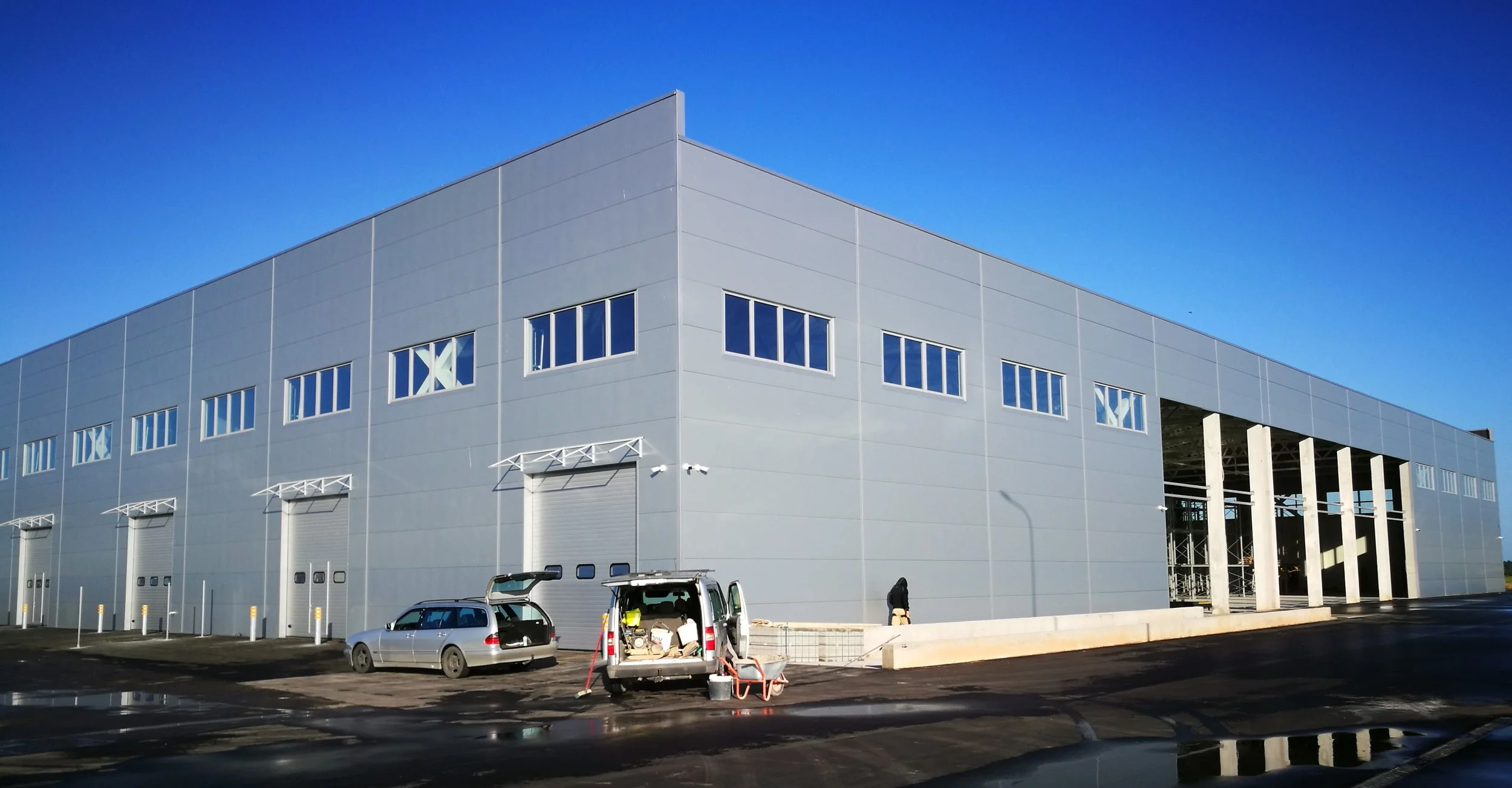 Large industrial warehouse building with multiple loading docks, parking lot with vehicles, and a person walking nearby on a clear day with blue sky.