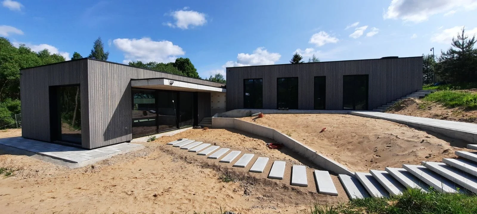 Modern house under construction with a wooden exterior, large glass windows, and a concrete pathway leading to the entrance, surrounded by a sandy yard and some greenery.