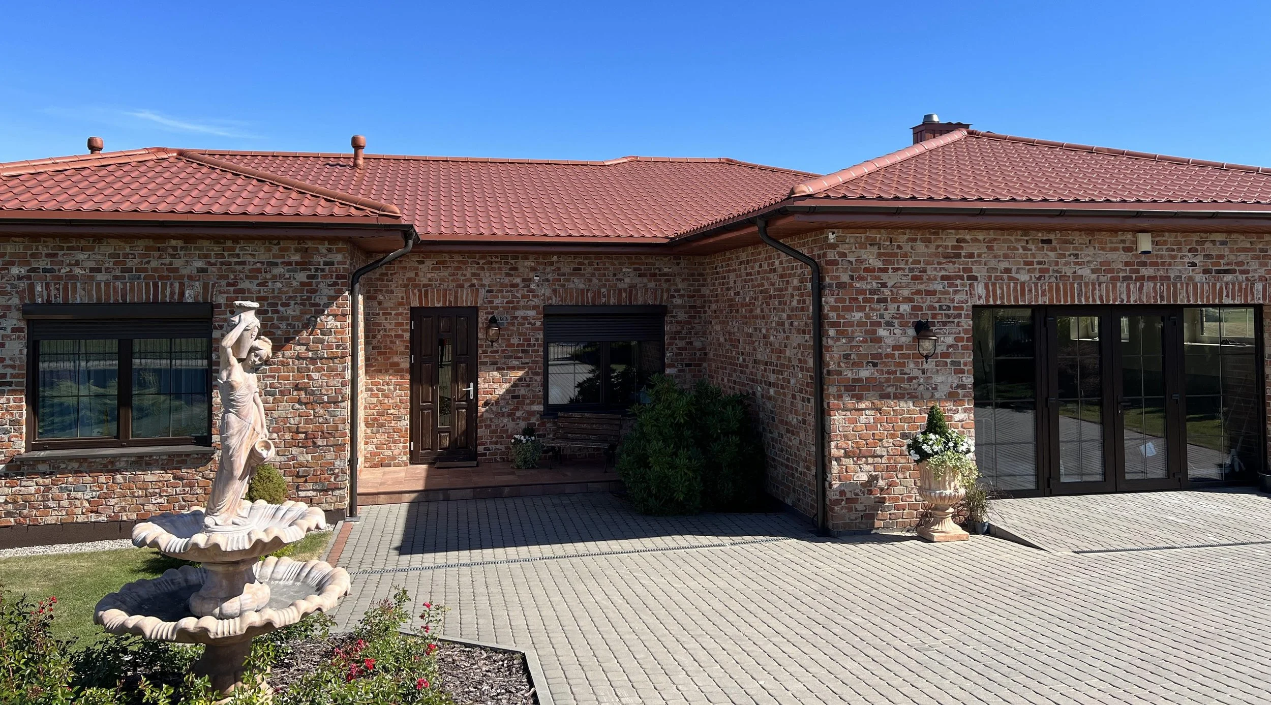 Front view of a brick house with a red tiled roof, black window frames, and a paved driveway, decorated with statues and planters.