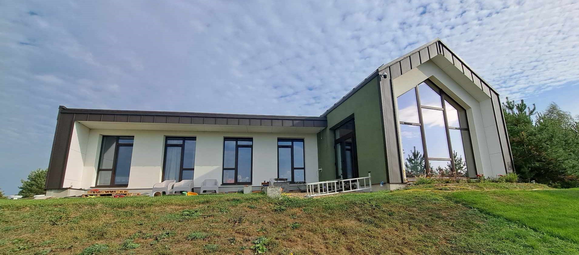 Modern house with large glass windows on a grassy hill, under a partly cloudy sky.