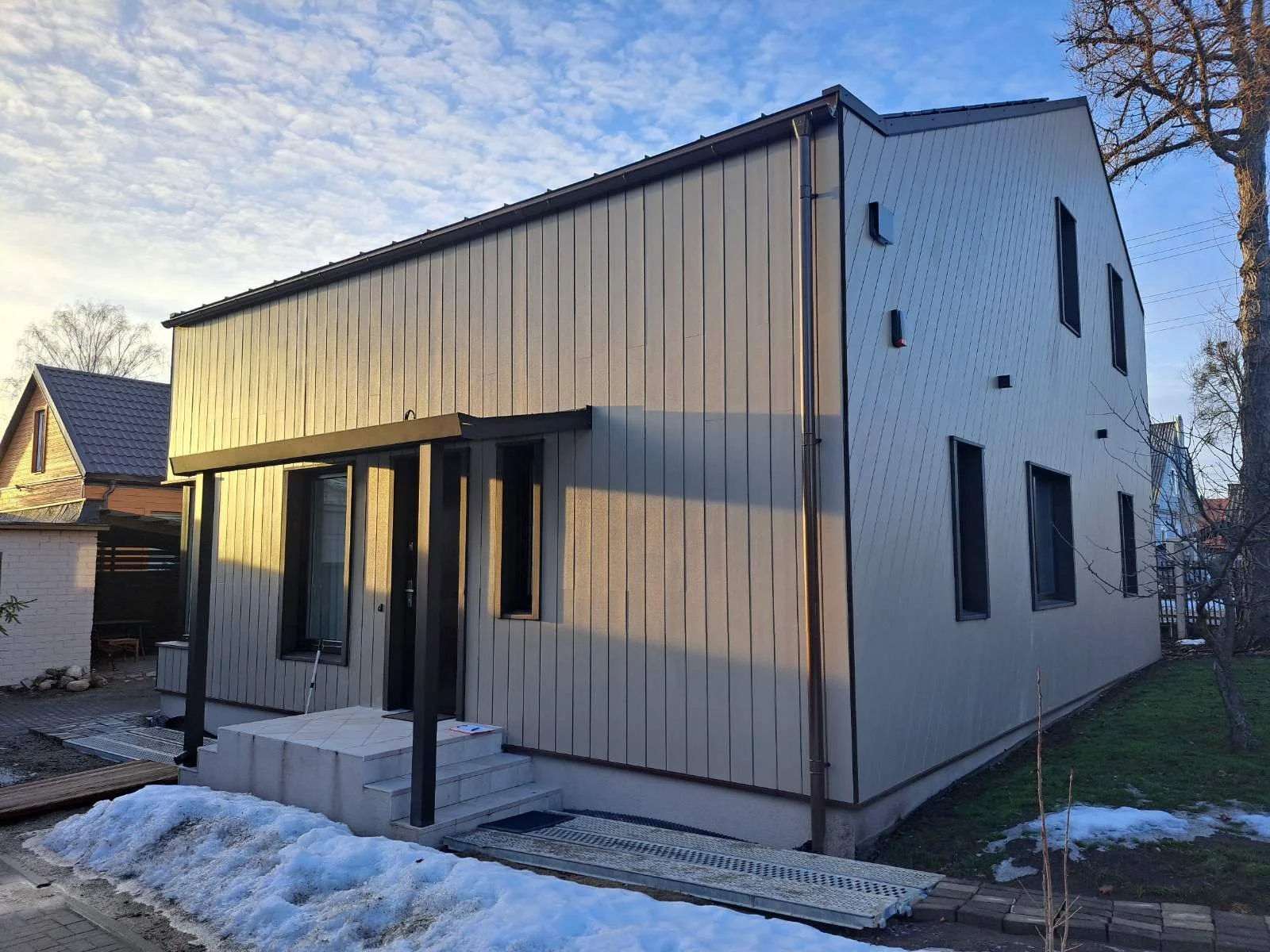 Modern two-story house with vertical beige siding and black window frames, small front steps, snow on the ground, trees, and neighboring houses