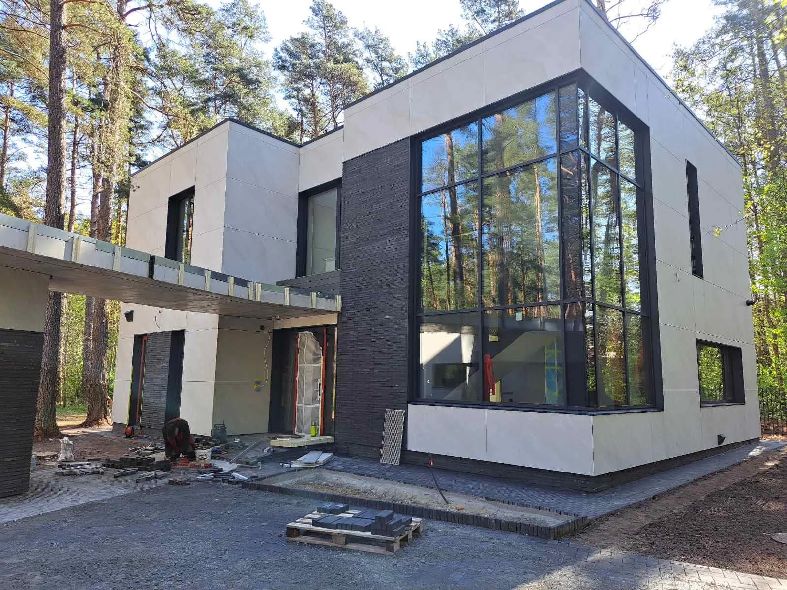 Modern house under construction surrounded by trees with large glass windows and white exterior panels.