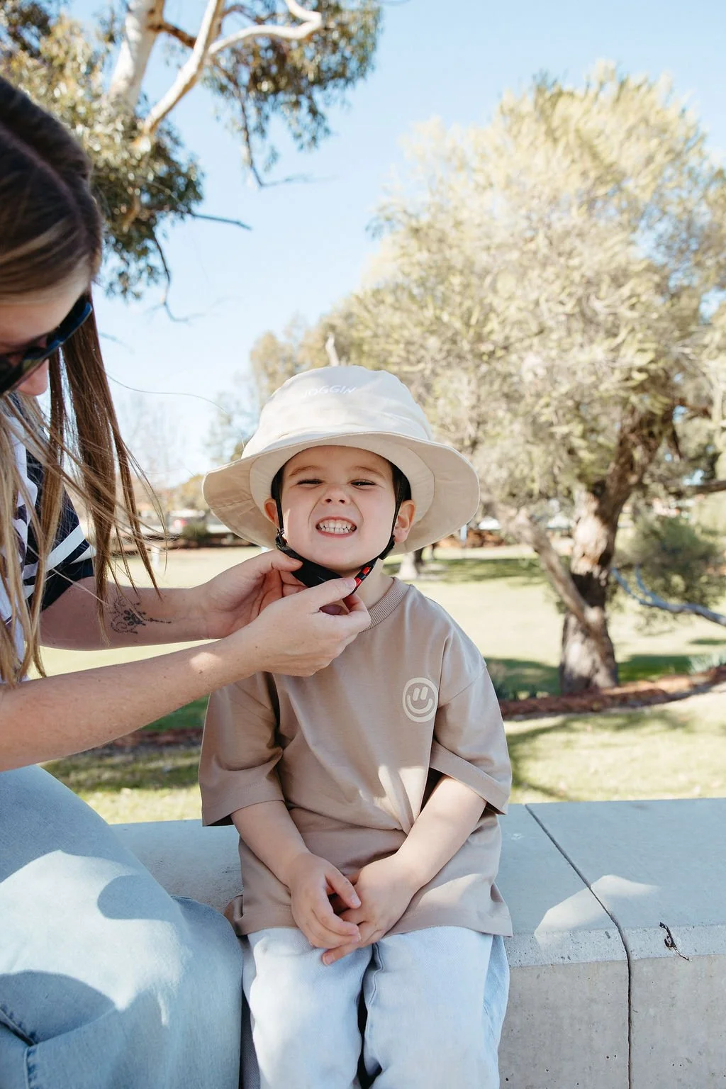 A young boy is sitting outdoors on a concrete bench, wearing a beige shirt and a wide-brimmed hat, with a playful expression showing his teeth. An adult woman is adjusting his black chin strap, with trees and a blue sky in the background.