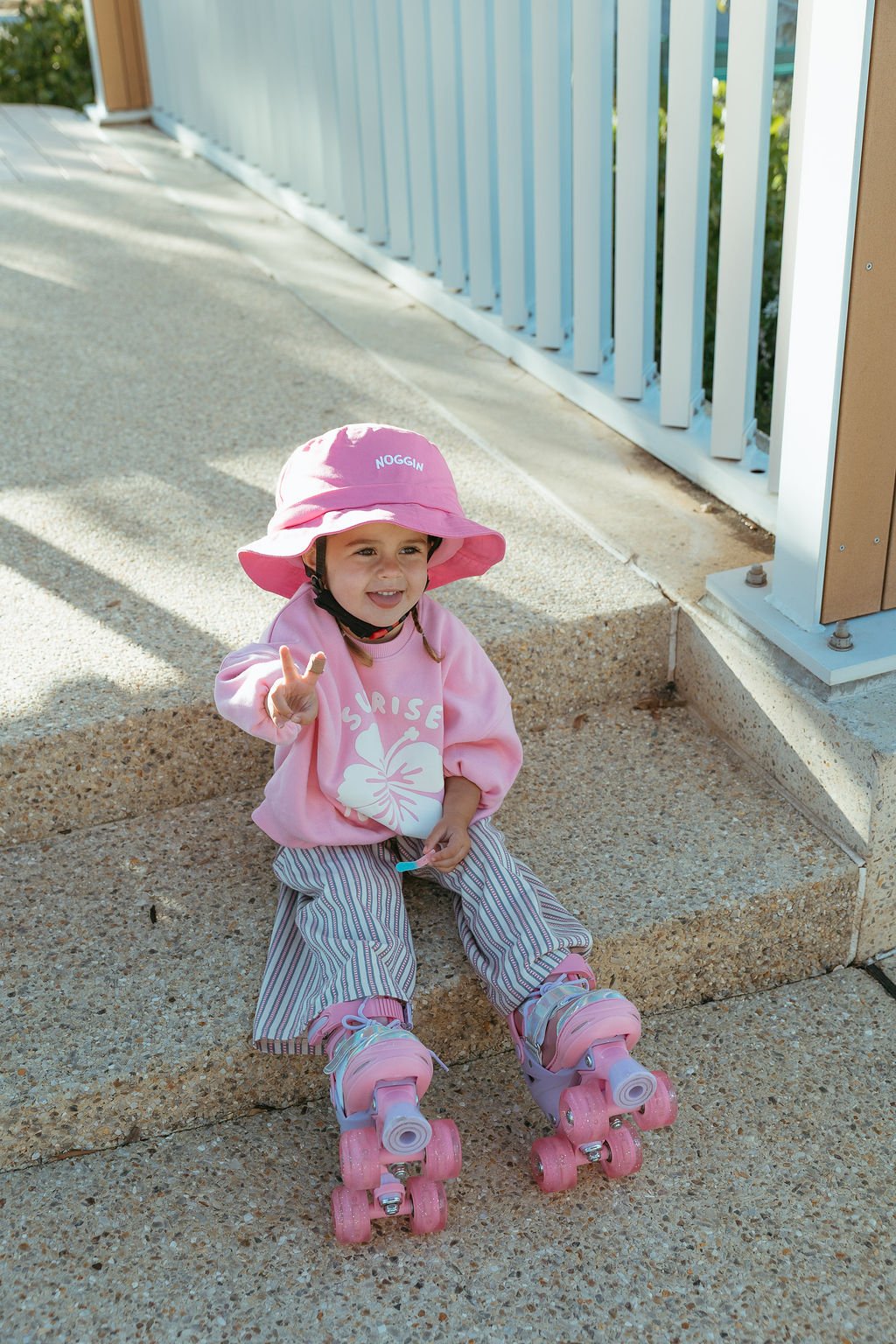 A young girl wearing a pink sun hat, pink sweatshirt, and striped pants sitting on a step with pink roller skates, making a peace sign and smiling.