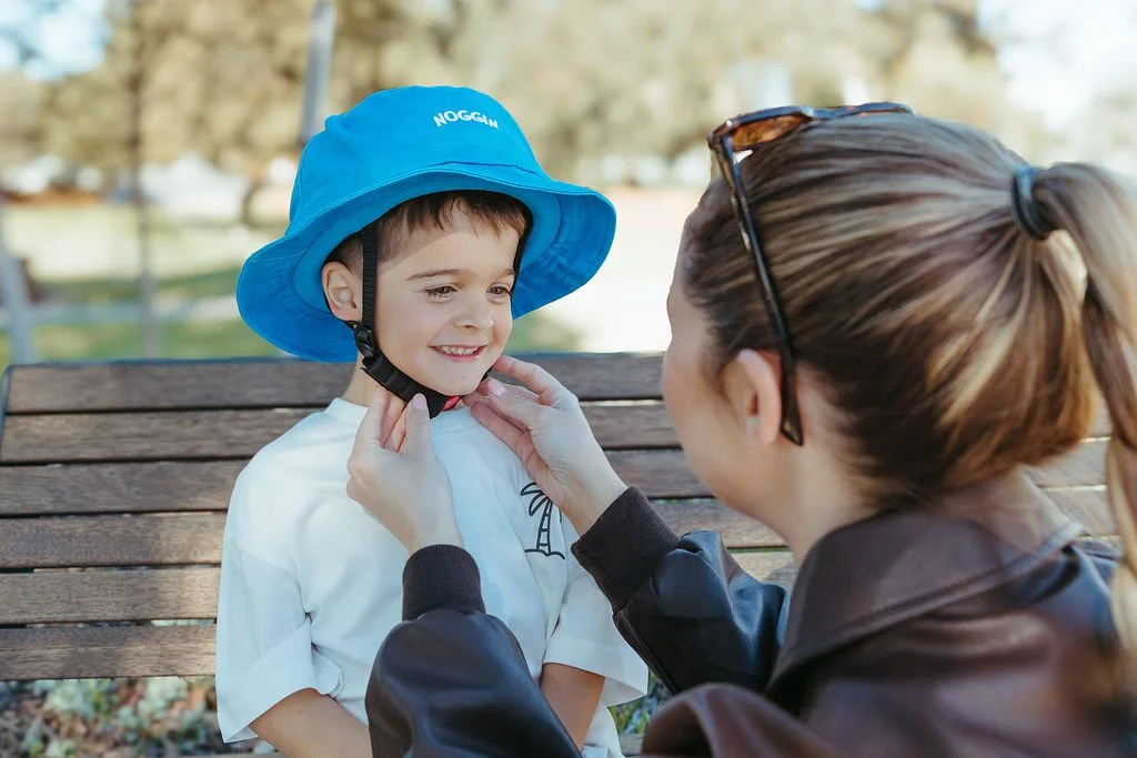 A woman helps a young boy put on a blue bucket hat while sitting on a park bench, during daytime with trees and water in the background.