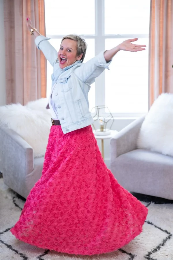 A woman joyfully dancing with her arms outstretched in a bright living room, wearing a pink textured maxi skirt and a light denim jacket.