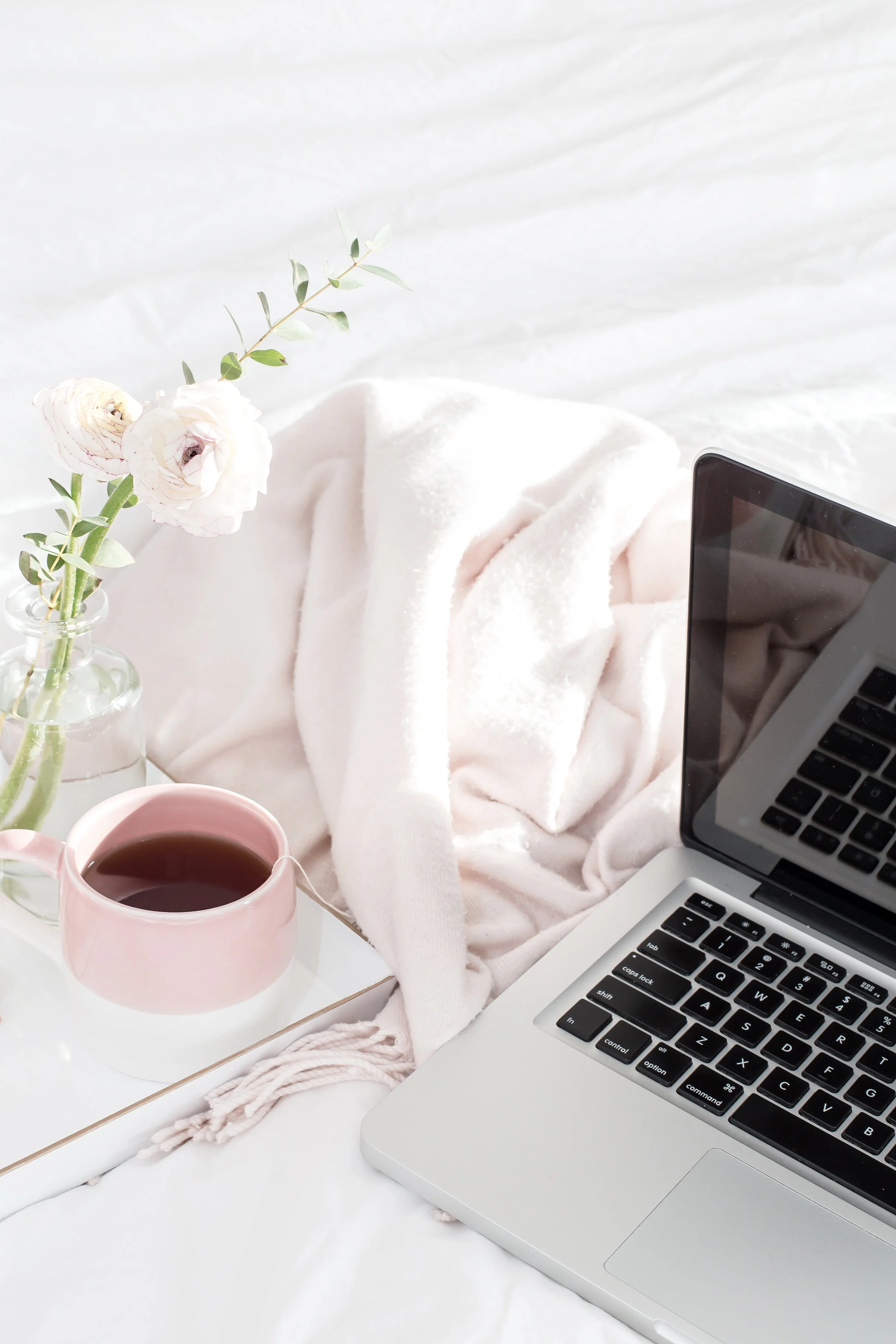 A cozy workspace with a silver laptop, a pink cup of tea, a vase with white flowers and eucalyptus, and a soft pink blanket on a white surface.