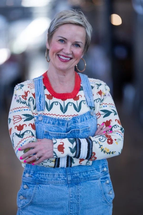 A woman with short blonde hair smiling, wearing a red and white patterned sweater, denim overalls, and large hoop earrings, standing outdoors with a blurred city background.