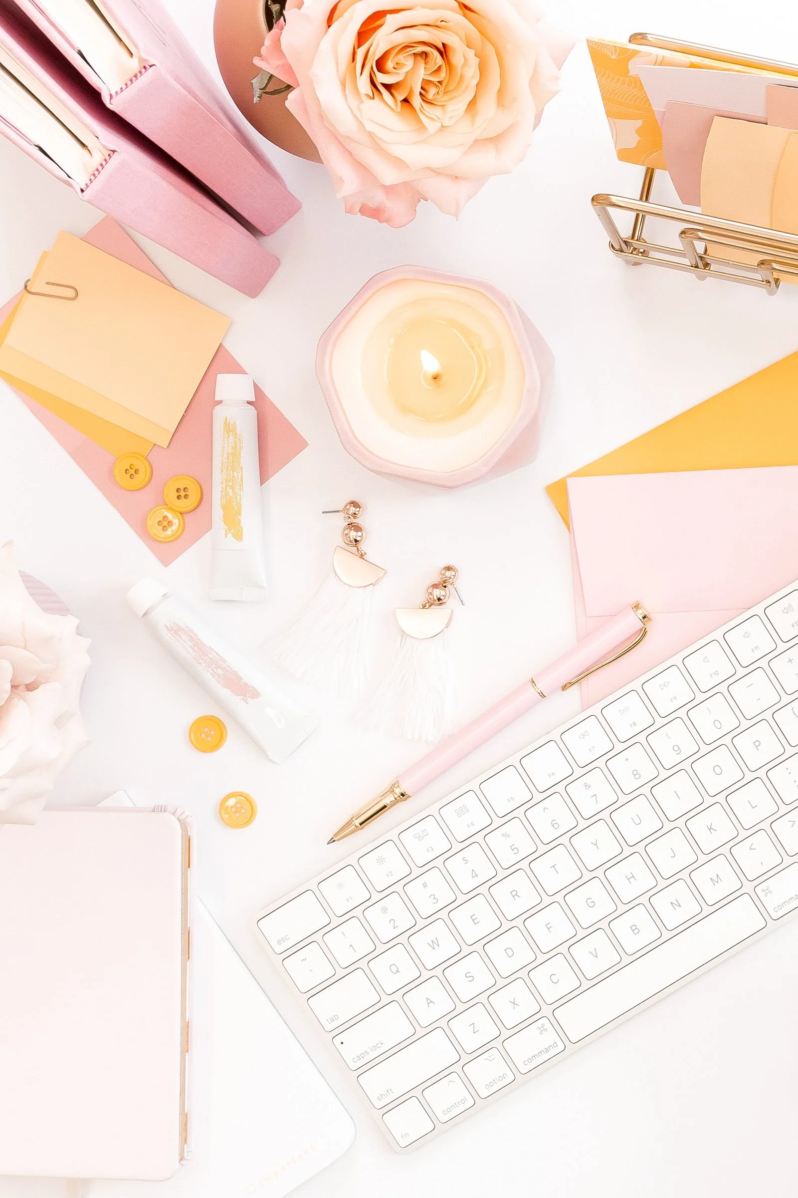 A pink and white workspace setup with a rose, a lit candle, a keyboard, a pen, earrings, folders, sticky notes, buttons, and a tube of paint on a white surface.