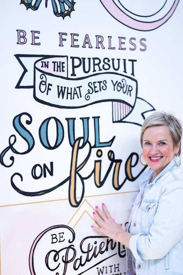 A smiling woman with short blonde hair wearing a light denim jacket standing next to a motivational sign with the quote, "Be fearless in the pursuit of what sets your soul on fire."