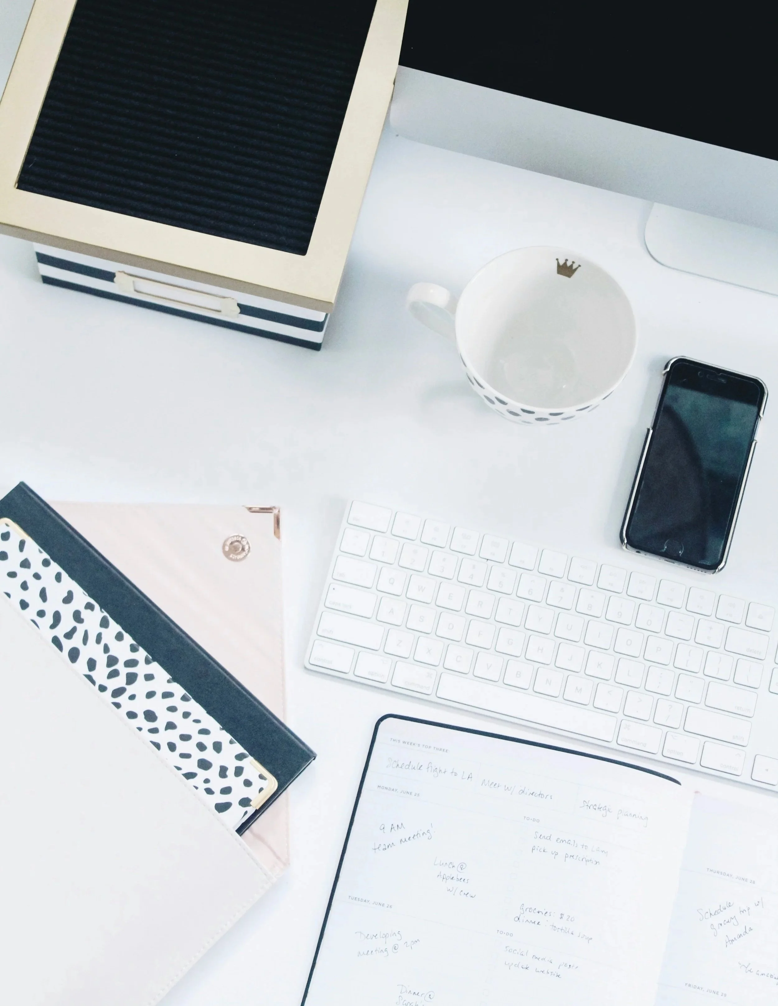 Desk with computer, keyboard, smartphone, a white coffee mug with a crown emblem, and papers with handwritten notes and a planner.