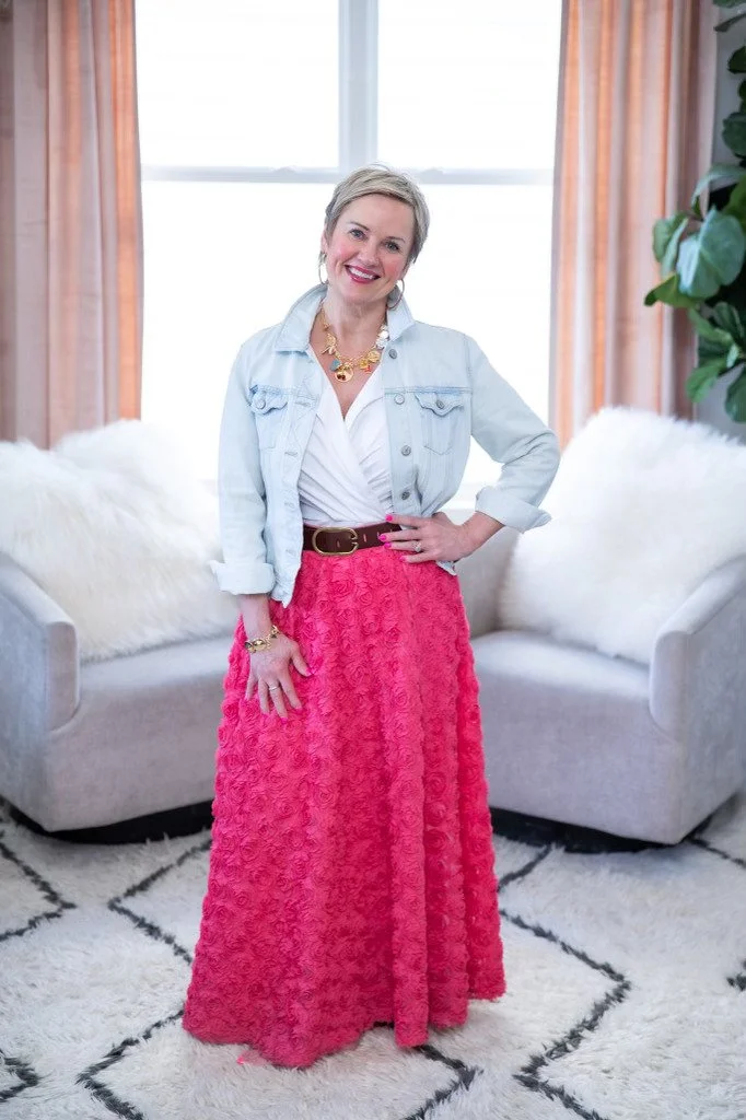 A woman with short gray hair smiling while standing in a brightly lit living room with peach curtains and green plants, wearing a denim jacket over a white blouse and a long pink textured skirt.