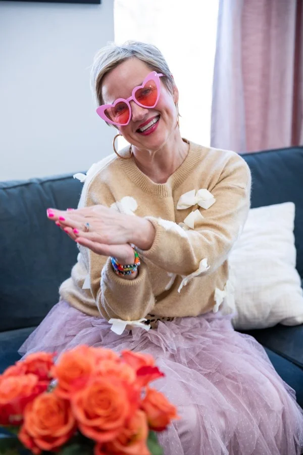 A smiling woman with short gray hair, pink heart-shaped sunglasses, wearing a beige sweater with bows and a pink tulle skirt, sitting on a black couch with cream pillows, clapping her hands with a bouquet of orange roses in front of her.