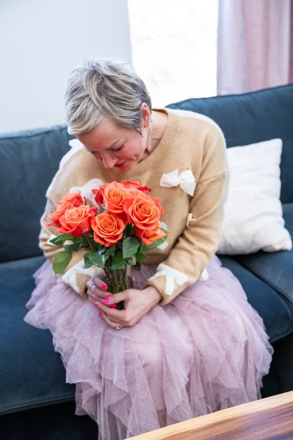 A woman sitting on a sofa holding a bouquet of orange roses, looking down at the flowers with a gentle smile.