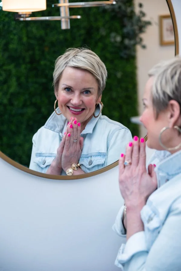 A woman with short blonde hair and pink nails looks at her reflection in a mirror, smiling with hands clasped near her face, in a room with a lush green wall background.