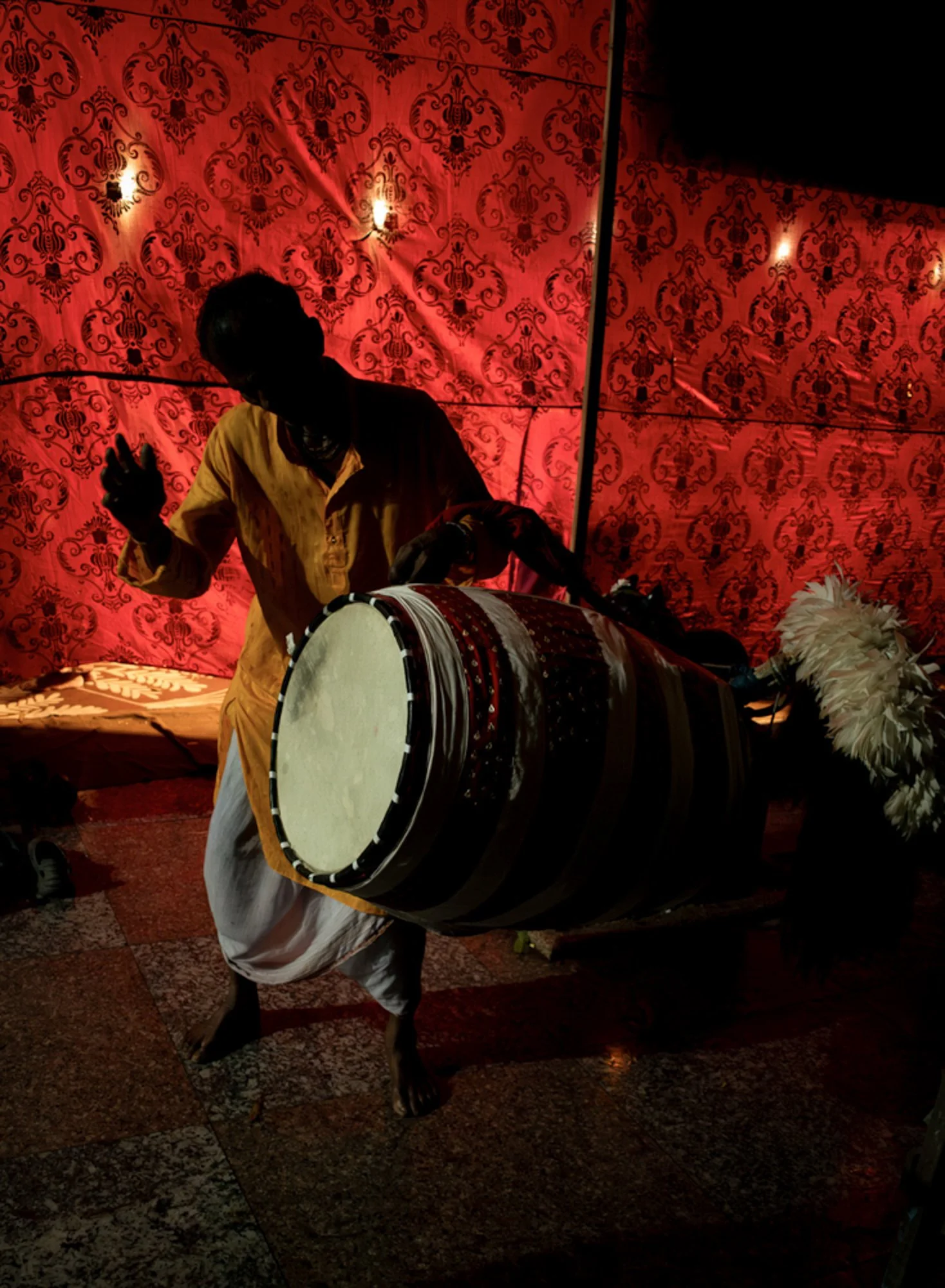 A person dressed in traditional Indian attire playing a large drum during a cultural performance, with a red patterned backdrop.