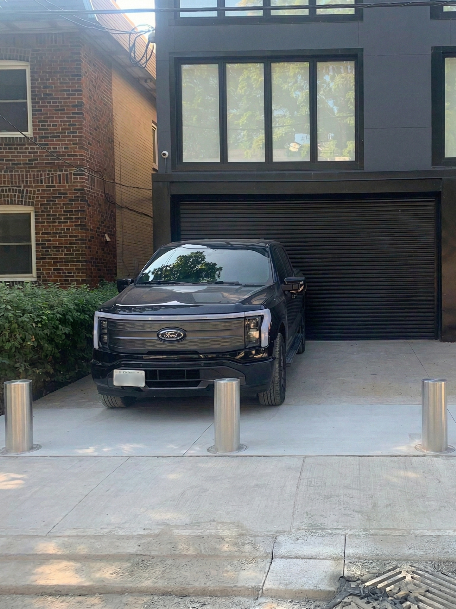 A black Ford truck parked in front of a modern building with large windows and black exterior, with a brick building on the left and small bushes on the left side of the parking spot, and metal bollards in the foreground.