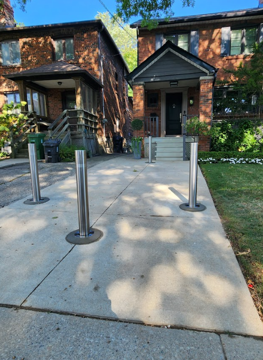 Three security bollards installed in a staggered pattern on a concrete driveway of a classic red brick Toronto house.