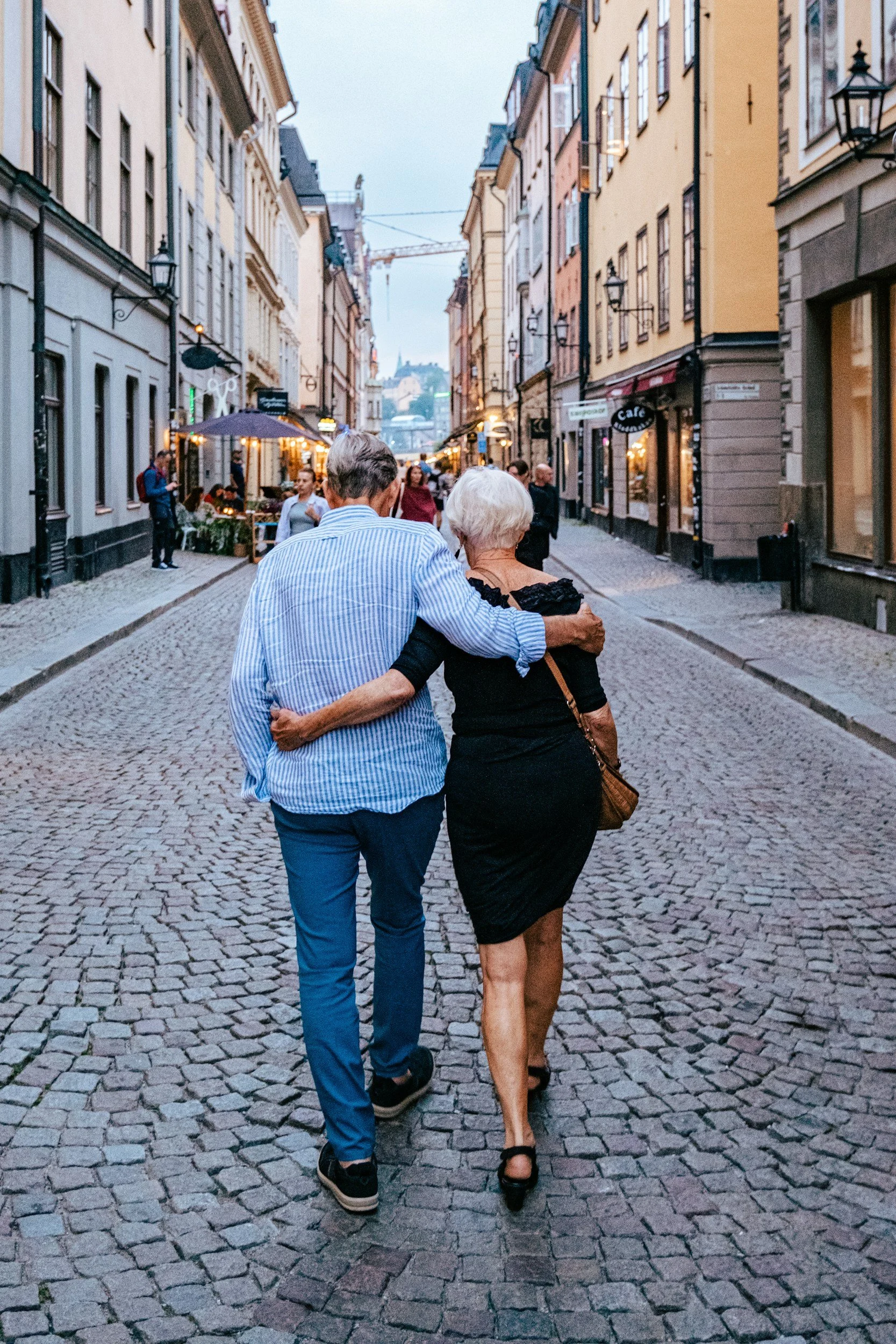 Ein älteres Paar geht Hand in Hand durch eine Kopfsteinpflasterstraße in einer Stadt. Im Hintergrund sind Gebäude mit geöffneten Geschäften und Lokalen zu sehen, sowie Straßenlaternen und eine Kräne. Es ist Abend oder Dämmerung.
