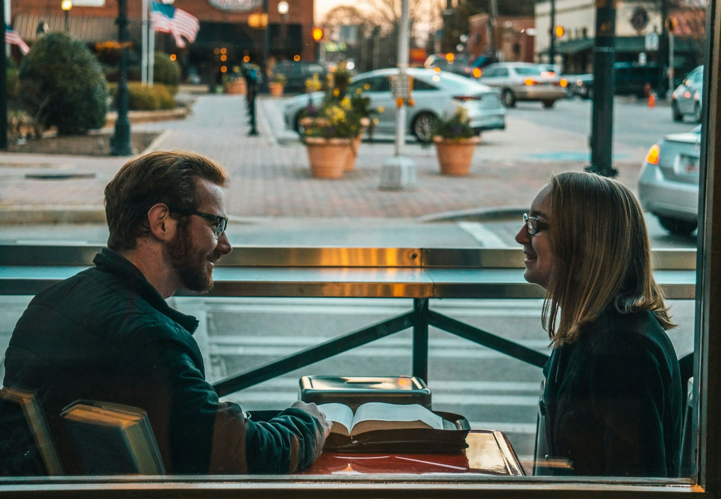 Zwei Menschen, ein Mann und eine Frau, sitzen sich gegenüber in einem Café, lachend und beim Lesen eines Buches oder einer Zeitschrift. Im Hintergrund sind ausgebliebene Autos und Pkws auf einer Straße samt Gehweg mit Pflanzen in Töpfen zu sehen.
