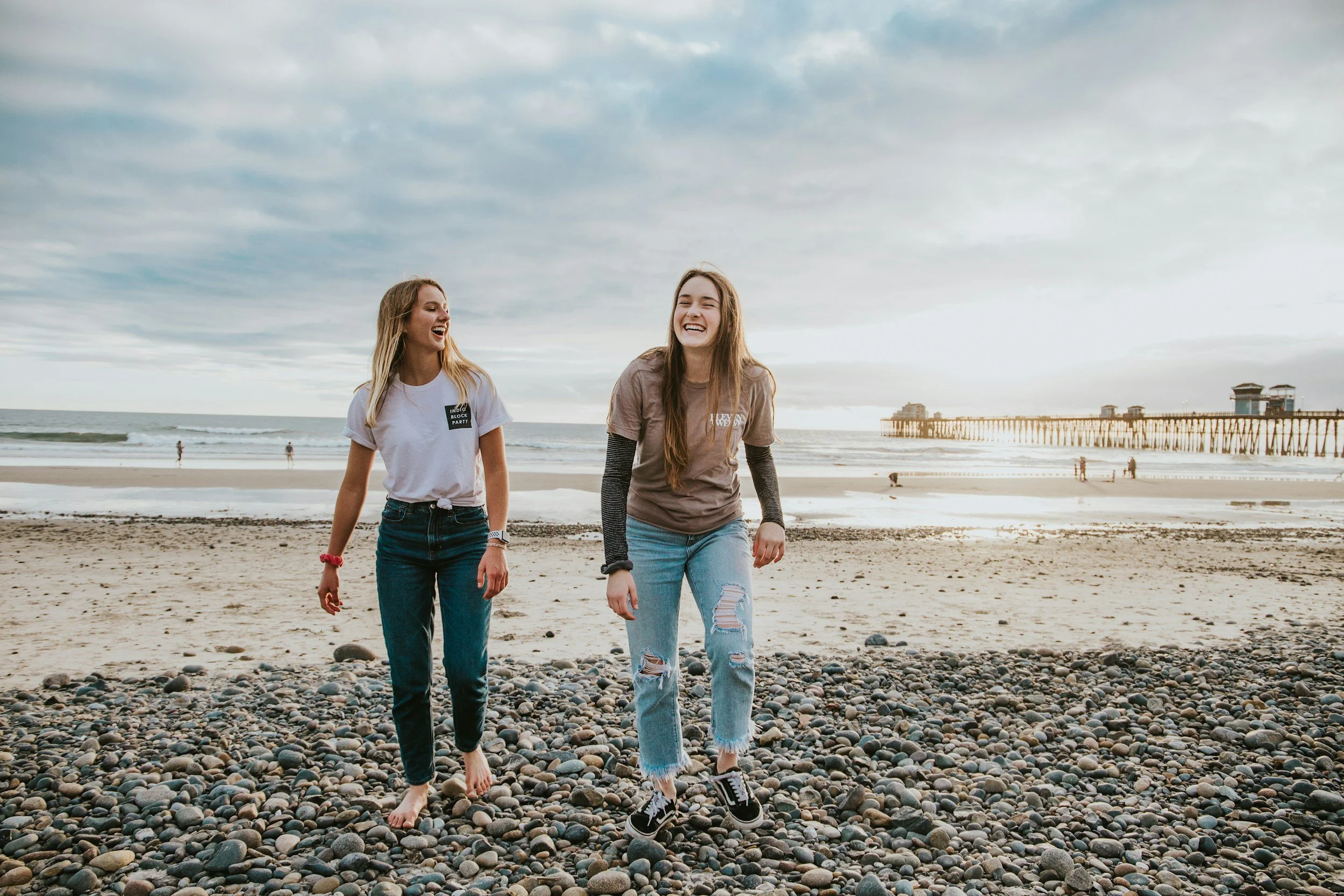 Zwei lachende junge Frauen gehen barfuß am Strand entlang, mit einer Pier im Hintergrund bei bewölktem Himmel.
